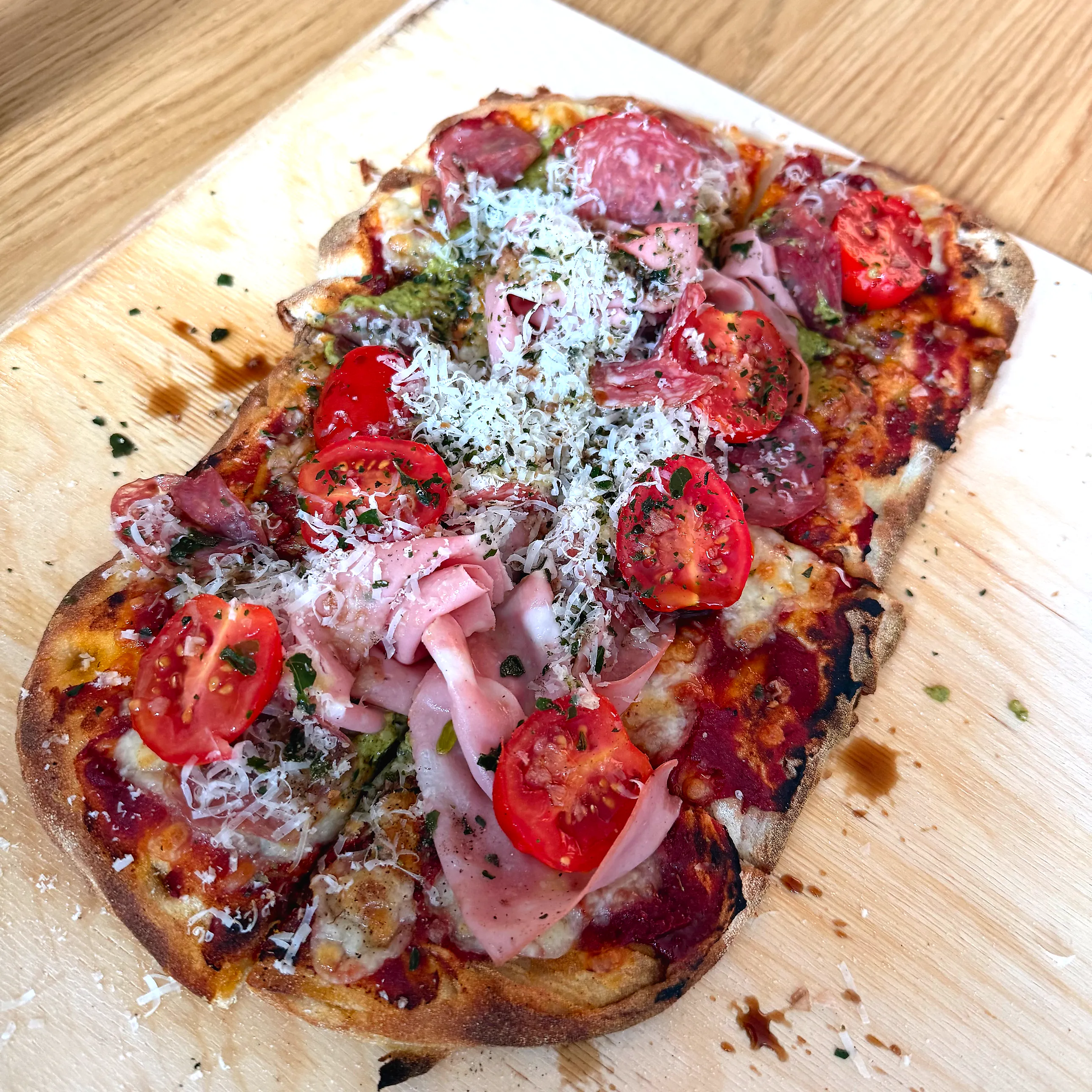 Avlång pinsa på träskärbräda med bubblig, mörkfläckig kant, täckt av mortadella, salami, halverade körsbärstomater, gröna pesto­klickar och ett tjockt lager finriven parmesan.