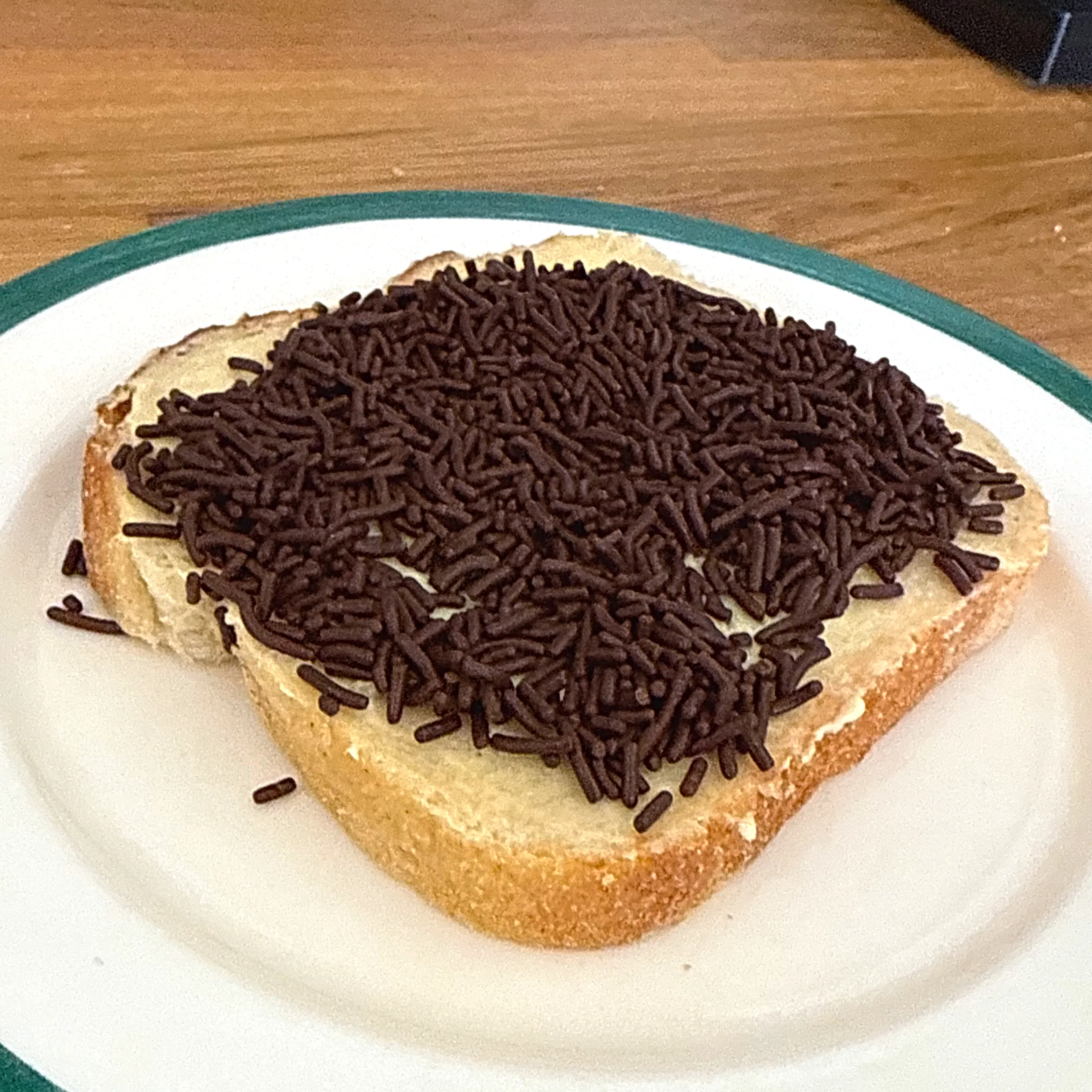 En skiva ljus formfranska med smör och ett tjockt lager mörkt chokladströssel, upplagd på en vit tallrik med grön kant på ett träbord.