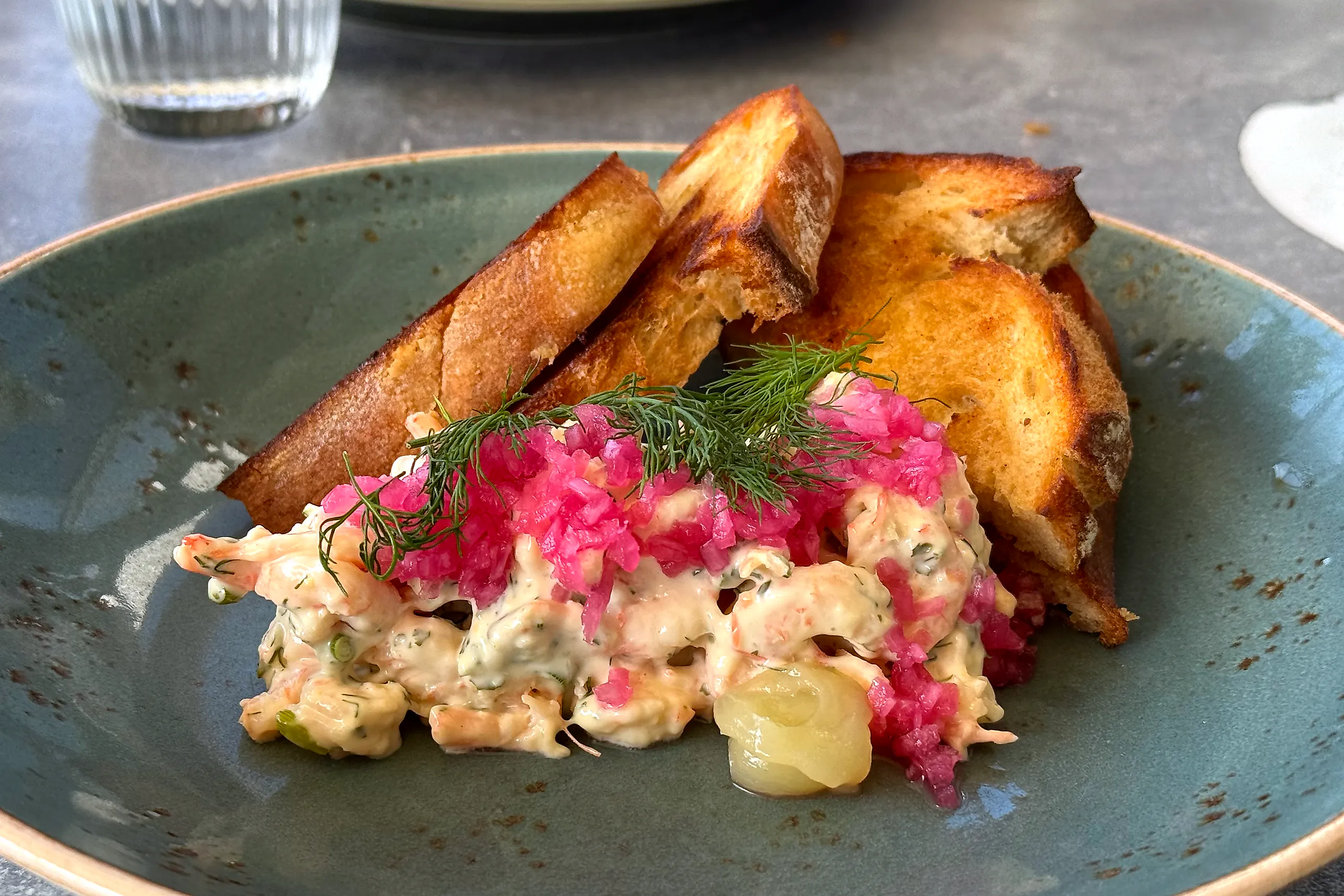 Krämig skagenröra med räkor och dill på en grön tallrik, toppad med starkt rosa picklad rödlök och dillkvistar, med gyllene rostade surdegsbrödskivor bakom.