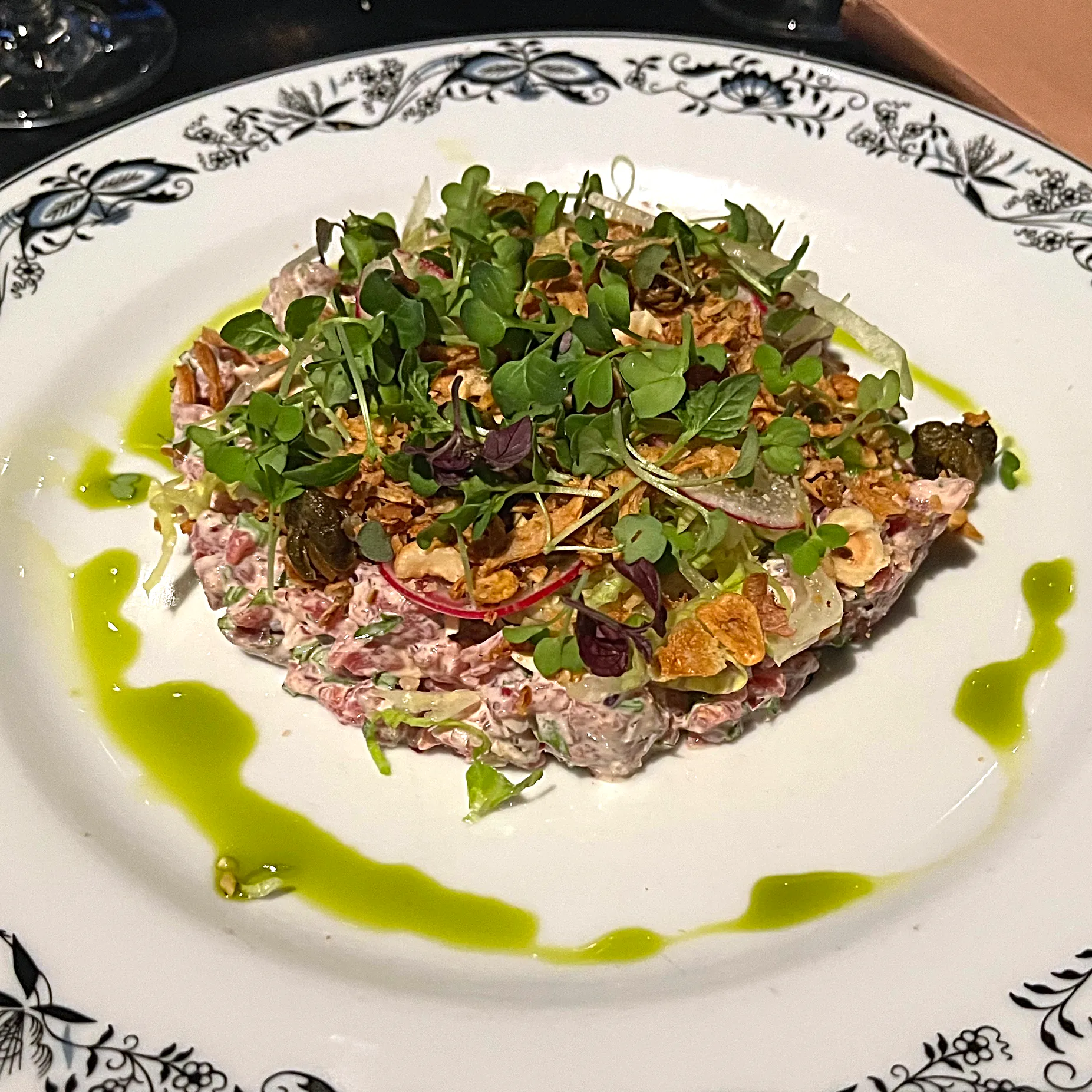 En rund kaka av krämig råbiff på en vitmönstrad tallrik, täckt av späda grönskott, frasiga lökchips, tunna rädisskivor, kapris och hasselnötter, med en ring av grön örtolja runt om.