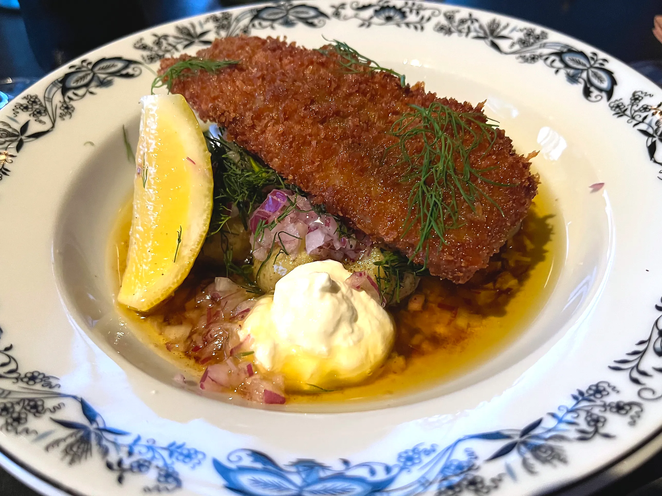 Djup tallrik med mönstrad kant där en gyllenbrun panerad fiskbit ligger på färskpotatis i brynt smör, med hackad rödlök, dillvippor, en klick vit crème och en citronklyfta.