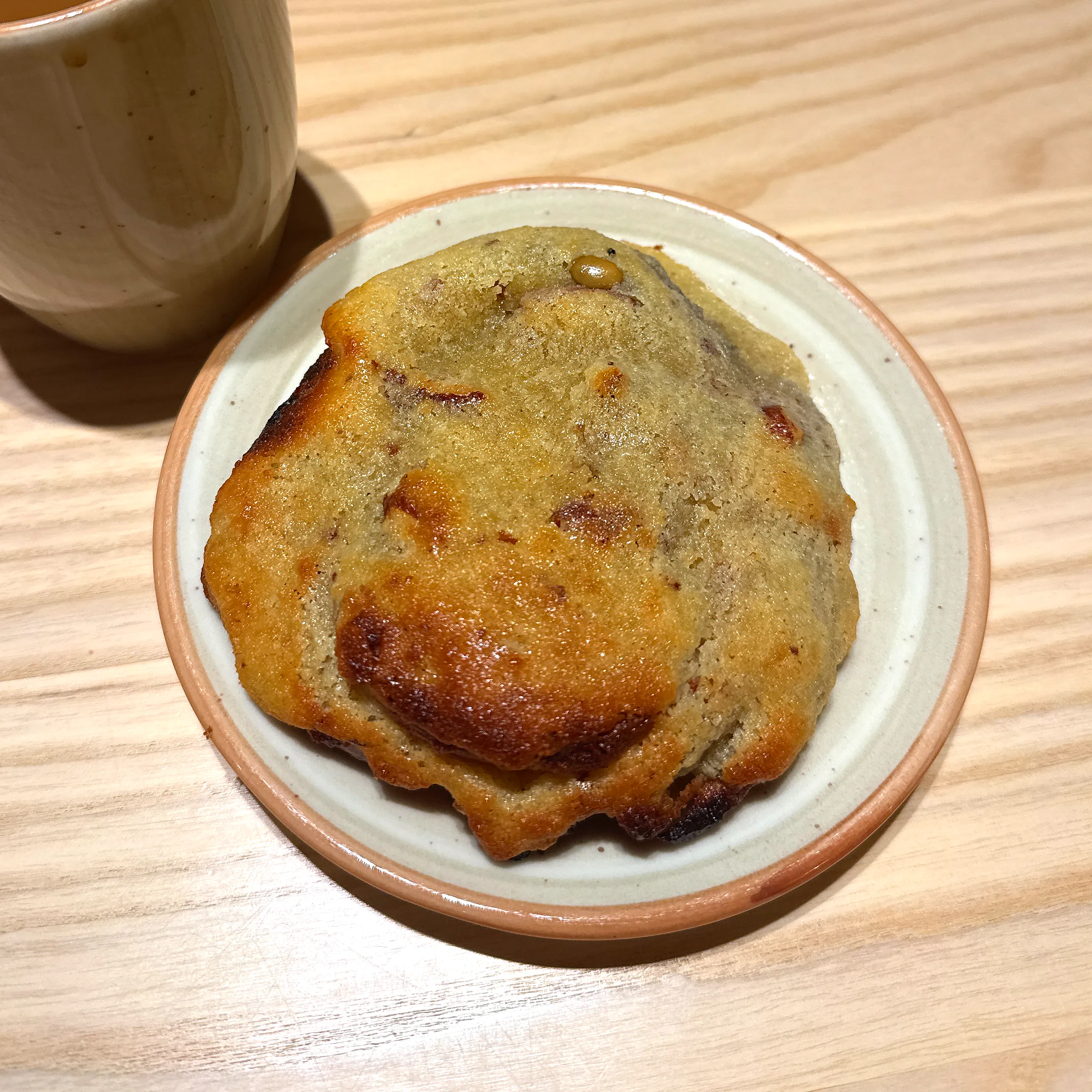 En stor, platt och gyllenbrun mandelbulle med ojämna, mörkbrynta kanter ligger på ett litet stengodsfat på ett ljust träbord, med en keramikmugg kaffe intill.