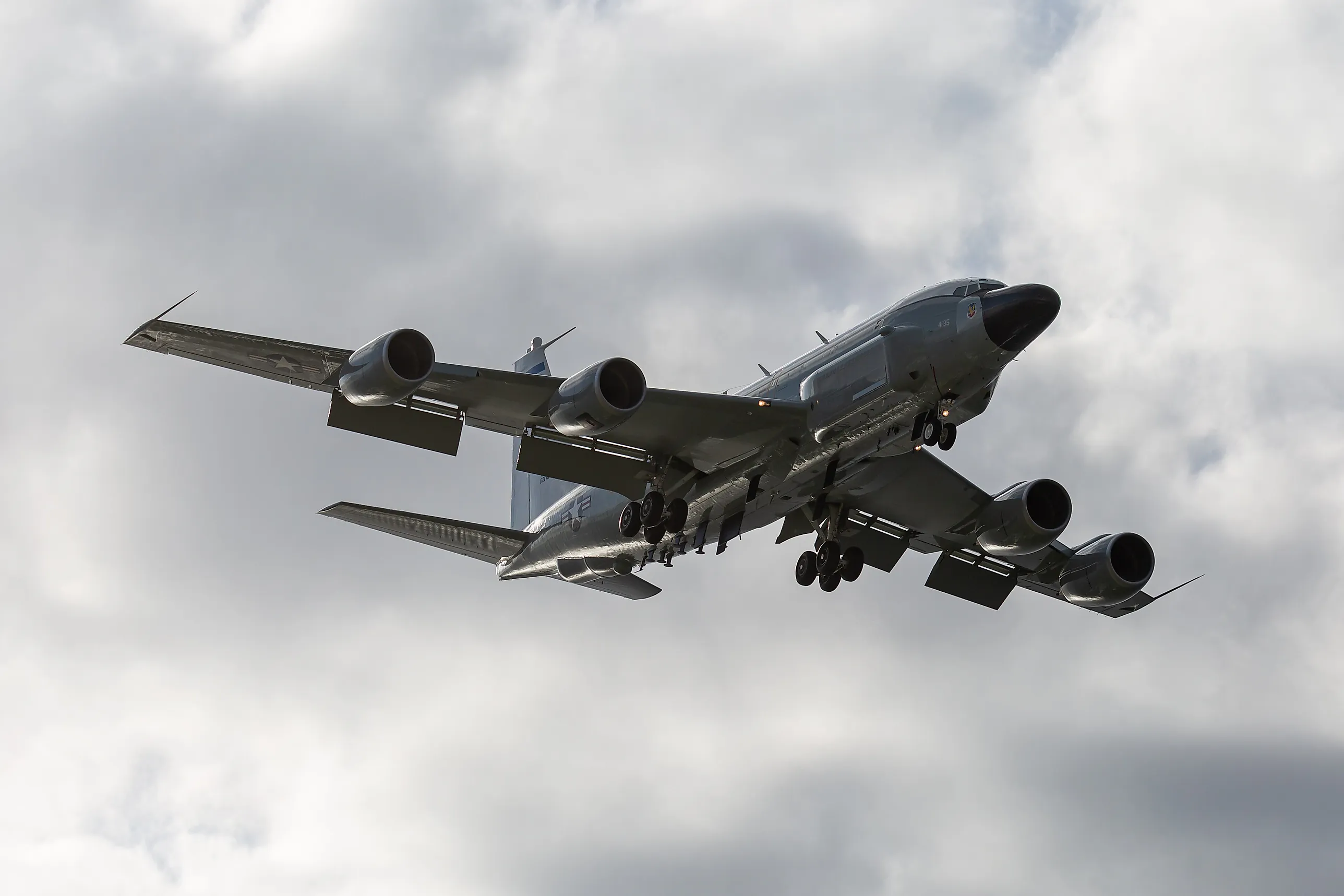 Ett grått fyrmotorigt spaningsplan av typen RC-135W med utfällt landningsställ, sett underifrån mot en molnig himmel.
