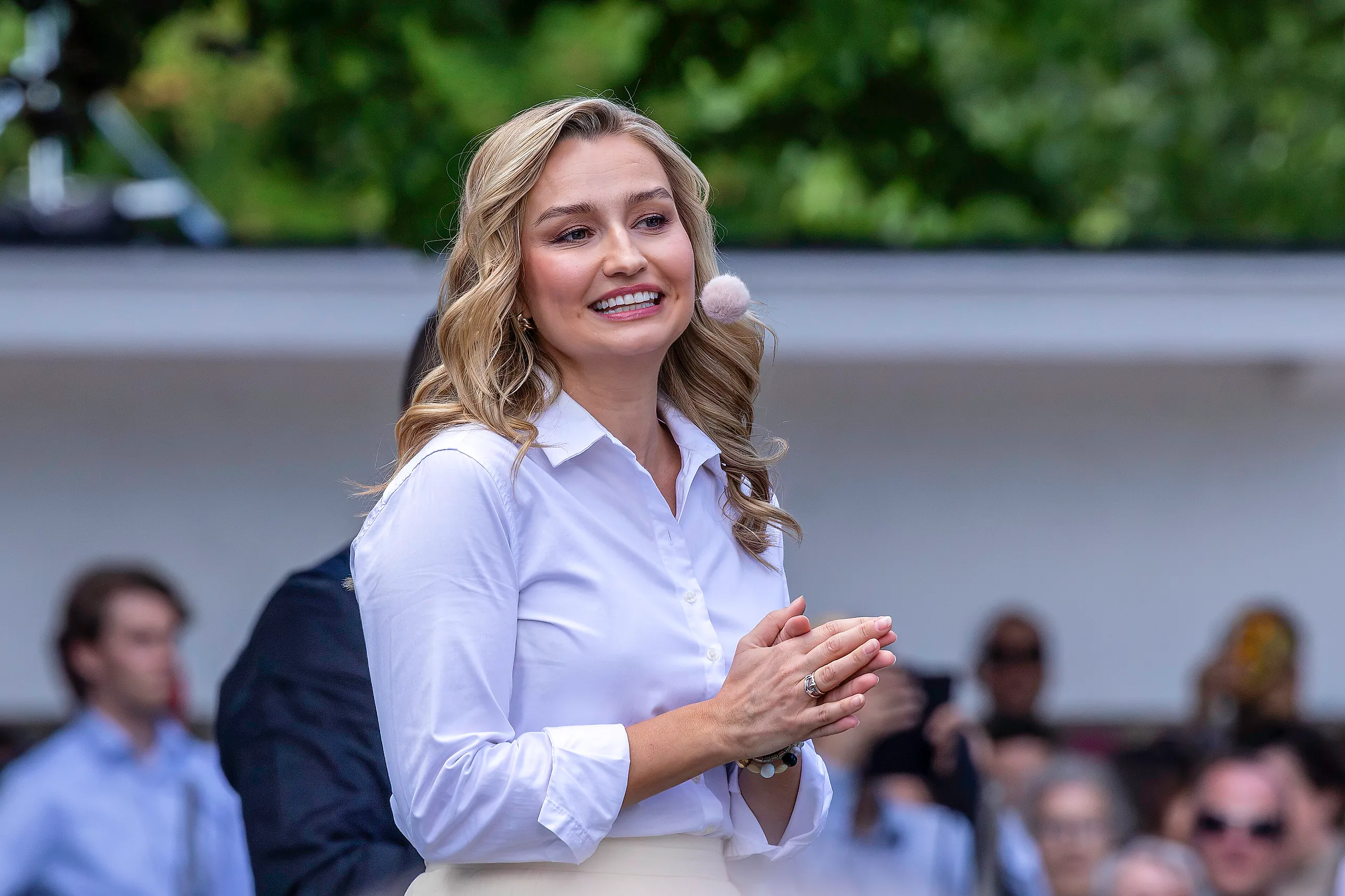 Ebba Busch i ljus skjorta står leende på en utomhusscen med händerna knäppta framför sig.
