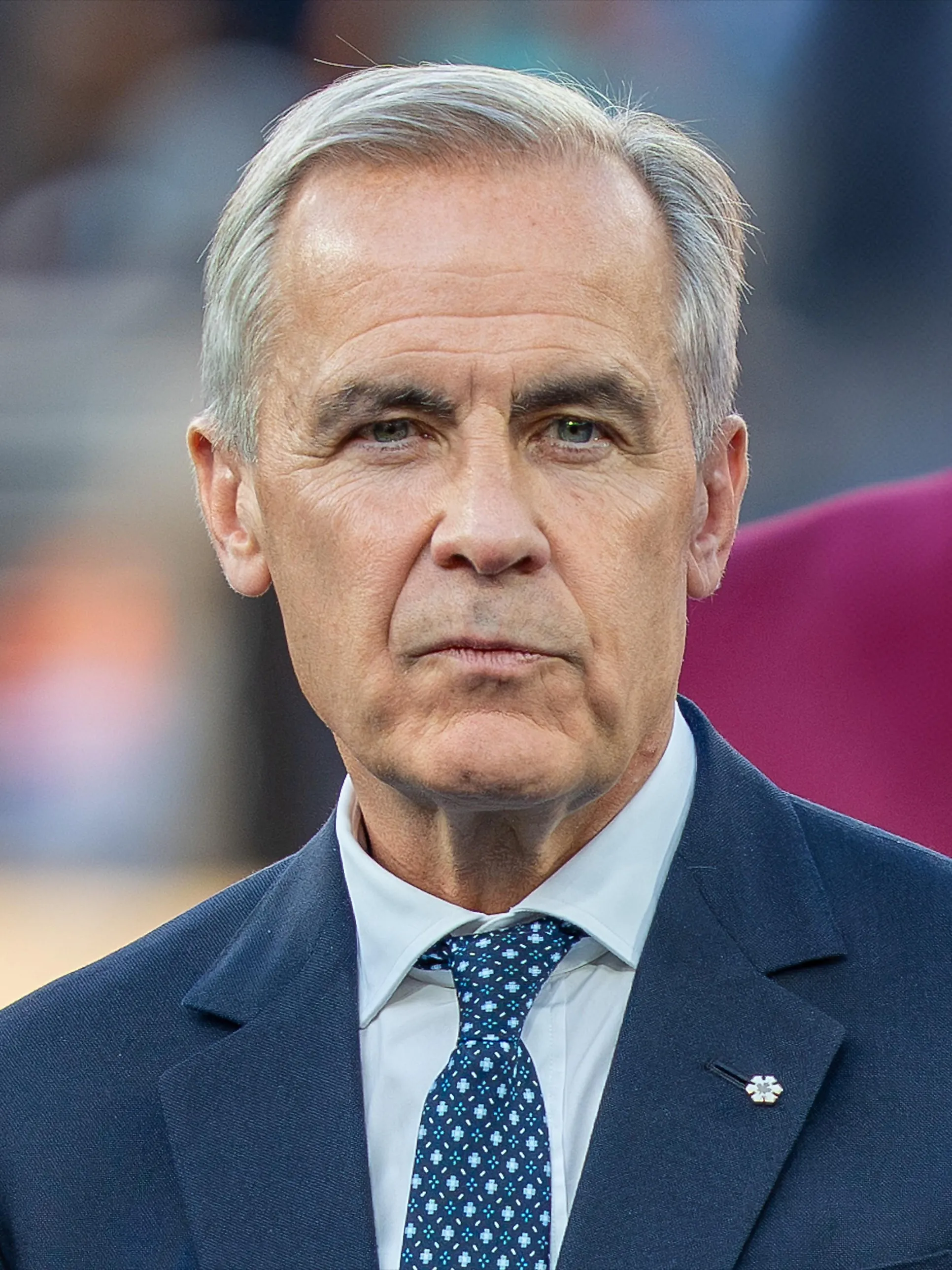 Mark Carney med allvarlig blick, i mörkblå kavaj med vit skjorta och mönstrad blå slips, mot en suddig bakgrund.