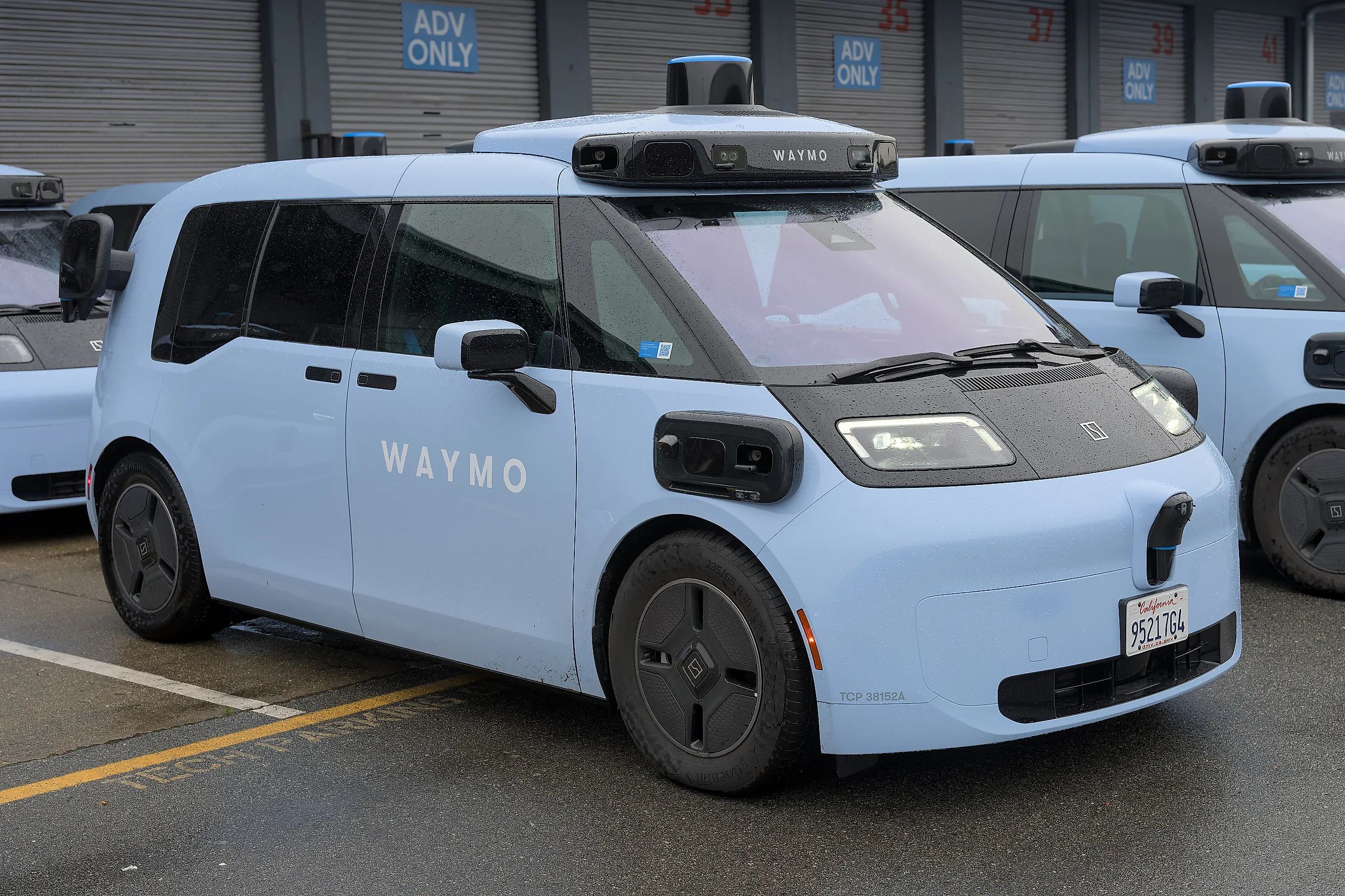 Ljusblå förarlös Waymo-taxi med sensortorn på taket parkerad framför en byggnad med portar märkta "ADV ONLY", med fler likadana bilar i rad bakom.