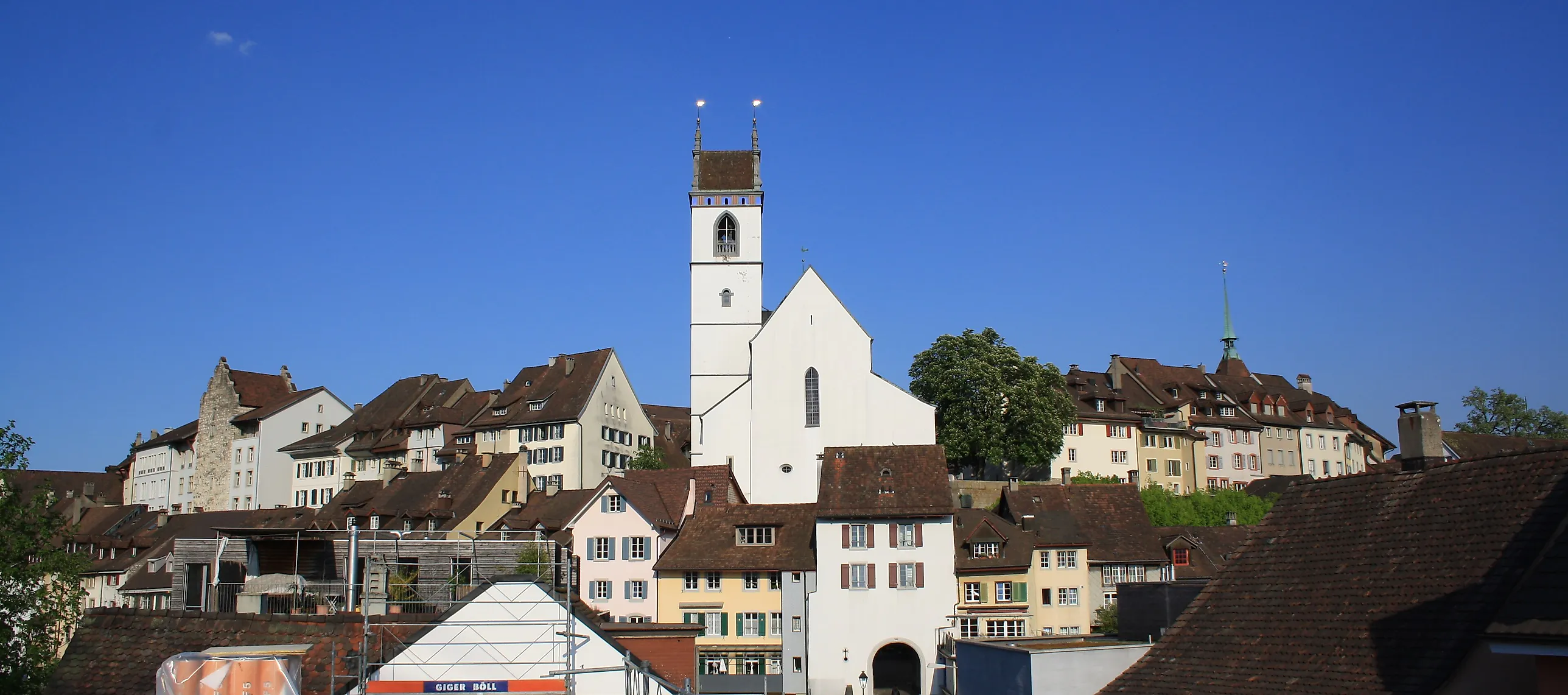 Gamla stadens tätt liggande hus med tegeltak och en vit kyrka med hög torn i Aarau under klarblå himmel.