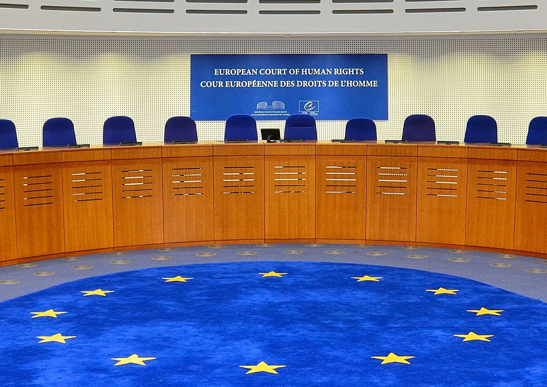 Tom rättssal med blå stolar bakom en böjd träbänk, skylten "European Court of Human Rights" på väggen och en blå matta med gula stjärnor på golvet.