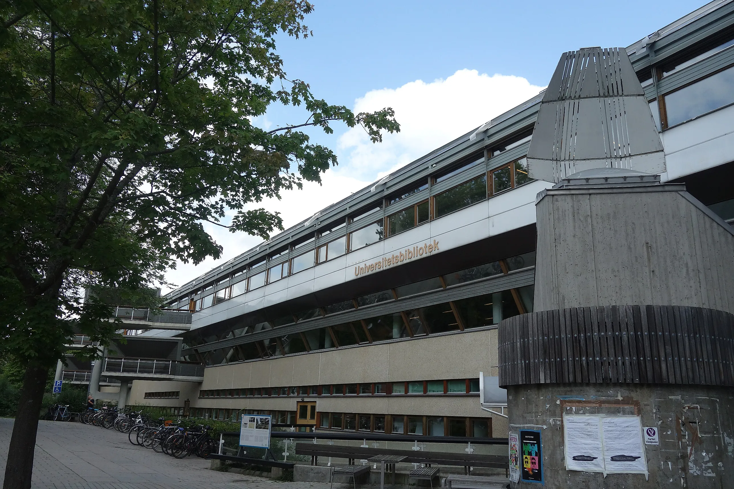 Betongbyggnad i flera våningar med skyltens text "Universitetsbibliotek" på fasaden, uppradade cyklar utanför entrén och ett lövträd till vänster i förgrunden.