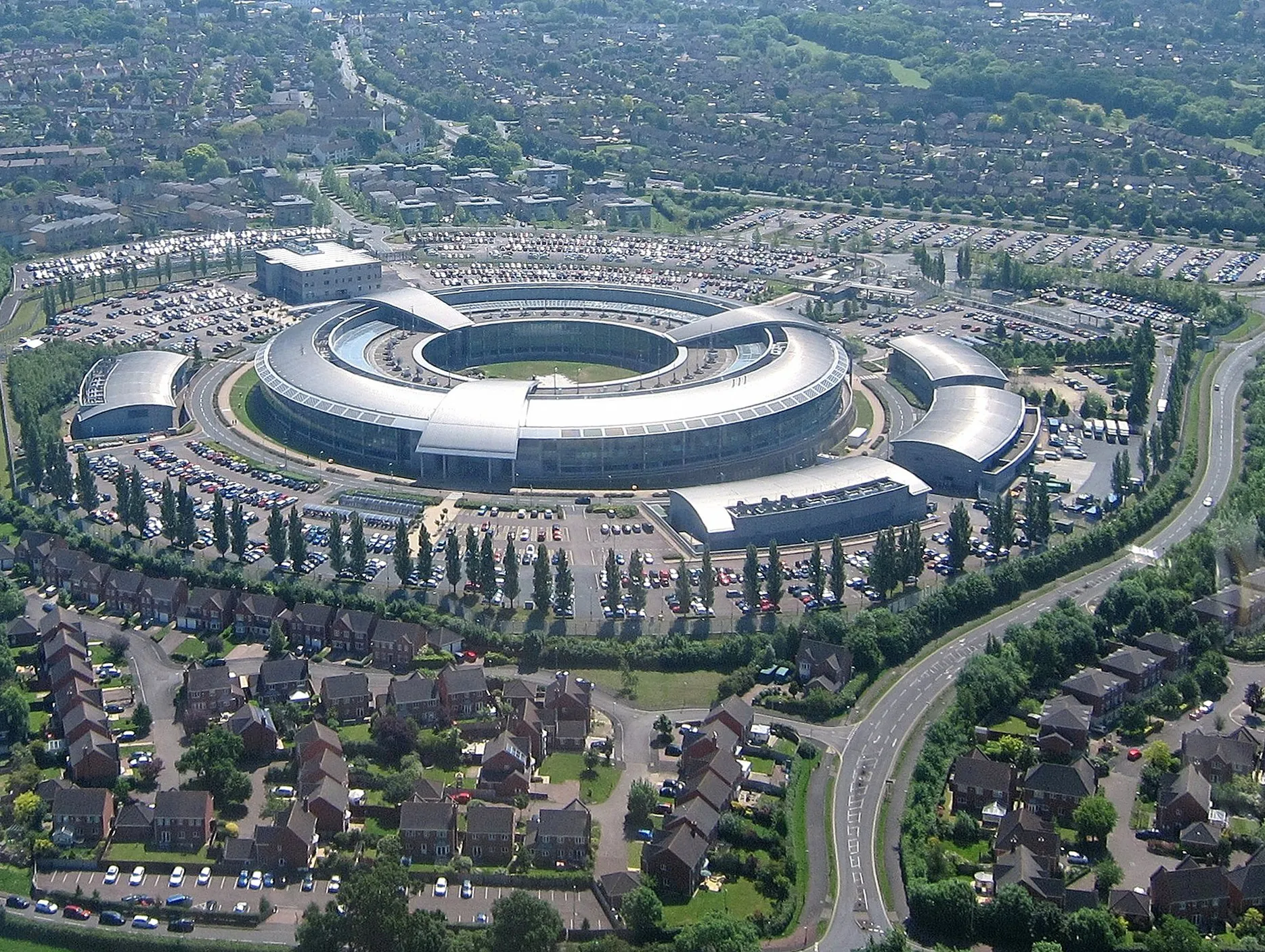 Flygvy över den ringformade byggnaden GCHQ i Cheltenham, omgiven av parkeringsplatser, trädrader och villakvarter.