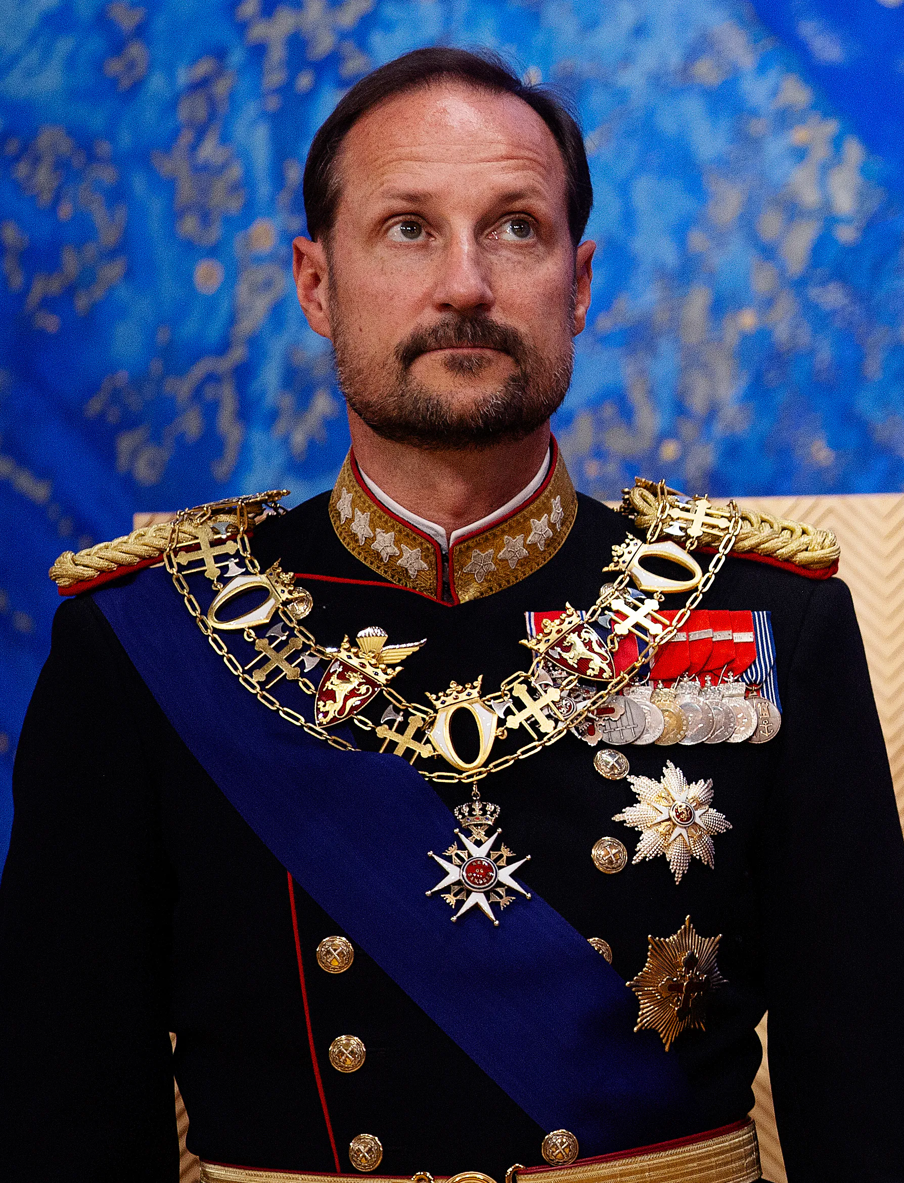 Haakon i mörkblå galauniform med guldepåletter, ordenskedja, blått axelband och rader av medaljer, mot en blåmönstrad vägg.