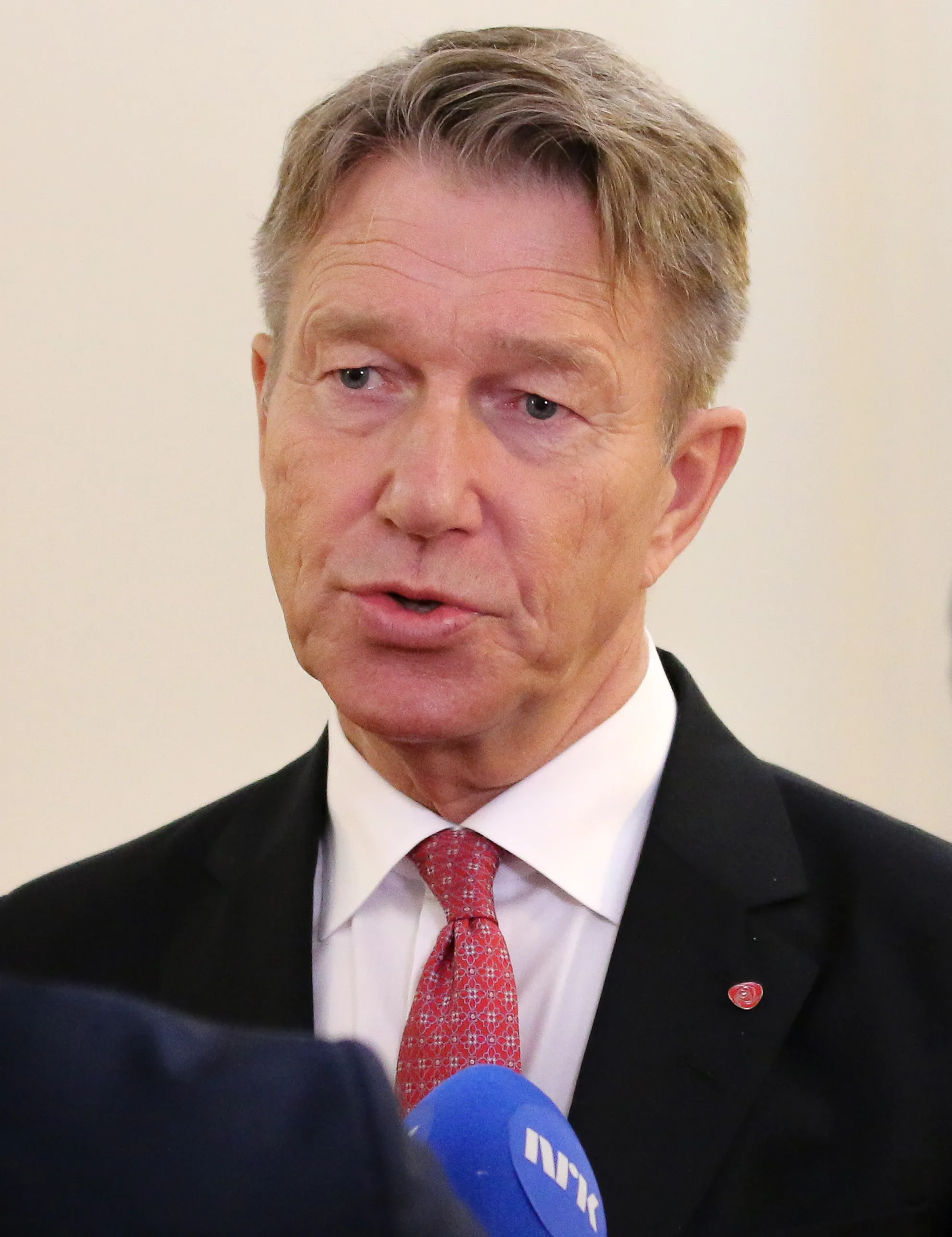 Terje Aasland i mörk kavaj och röd mönstrad slips talar till en blå mikrofon märkt NRK som syns i nedre bildkanten.