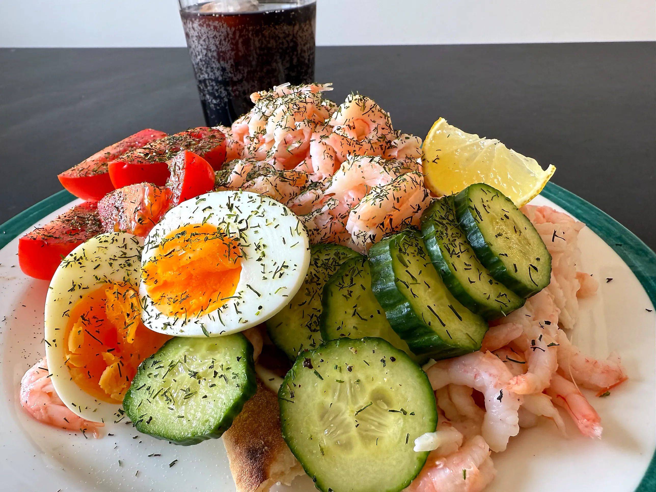 Tallrik med hönökaka täckt av handskalade räkor, halverat ägg med krämig gula, tjocka gurkskivor, tomatklyftor och en citronklyfta, allt strött med dill.
