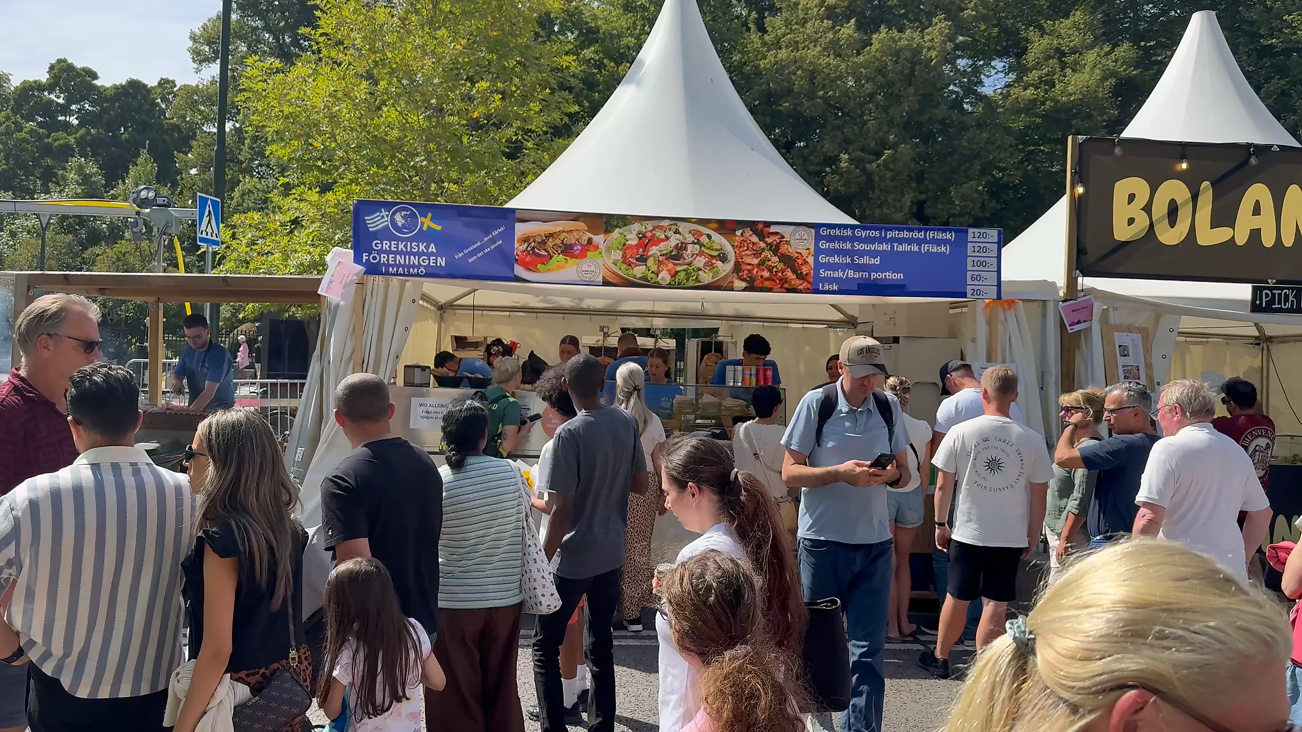 Kö av besökare framför ett matstånd med skylten Grekiska Föreningen i Malmö och prislista på gyros och souvlaki, med vita festivaltält bakom.
