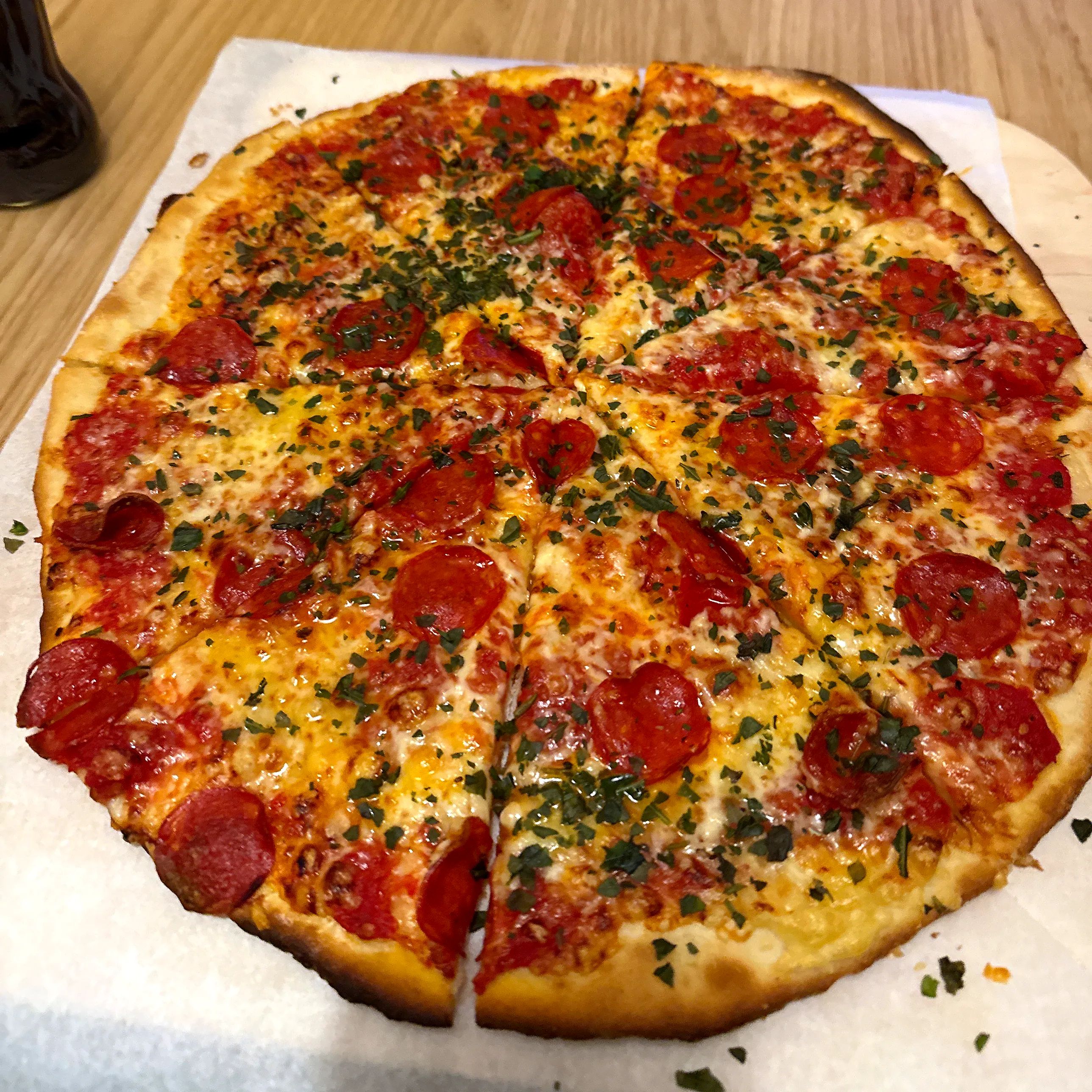 Rund, tunn pizza med tomatsås, smält ost och pepperoniskivor, rikligt strödd med hackad persilja, uppskuren i bitar på vitt papper på en träbordsskiva.