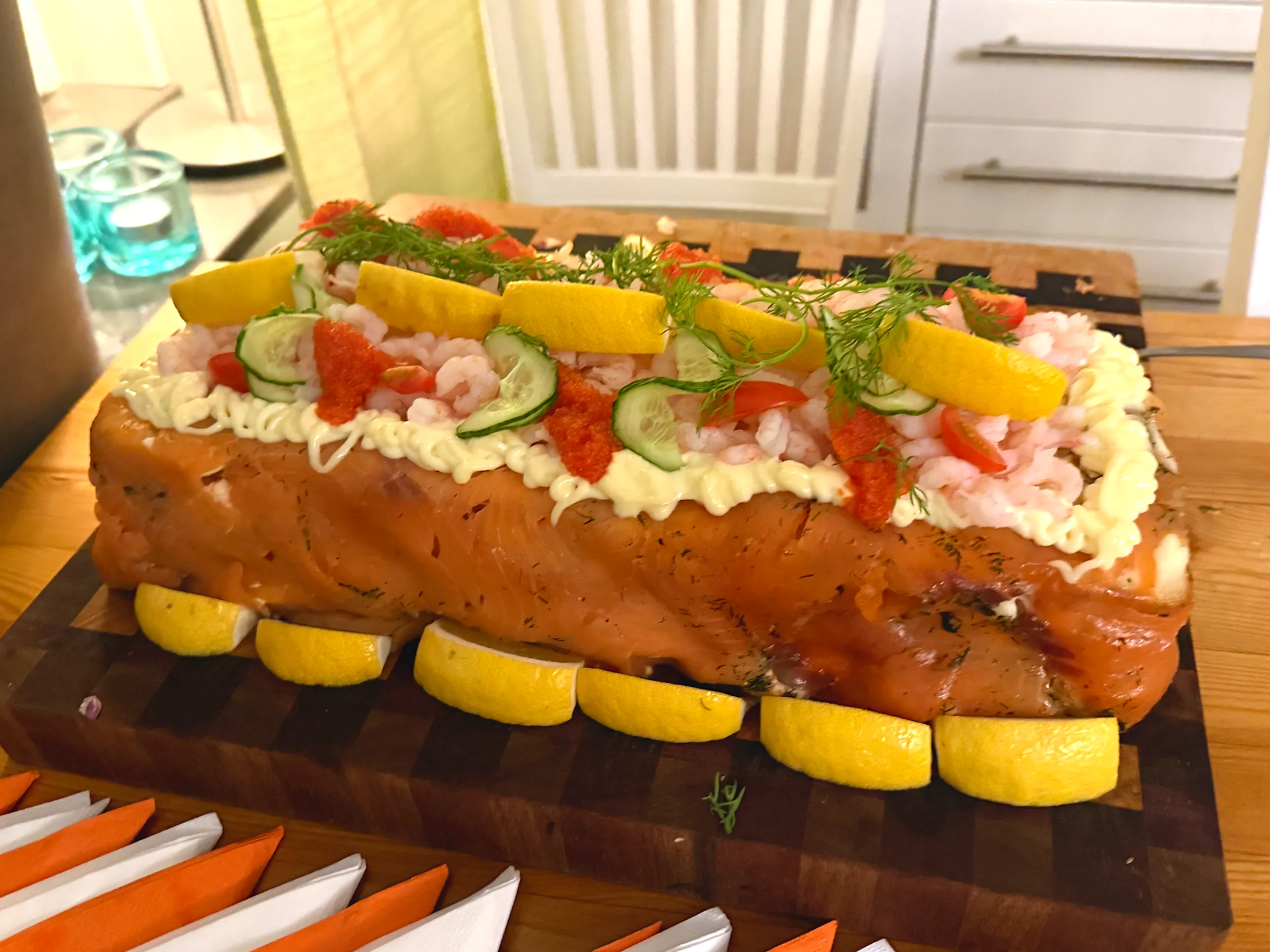Rektangulär smörgåstårta klädd i rökt lax, med bård av spritsad majonnäs, räkor, löjrom, gurkskivor, tomat, citronskivor och dill på toppen, omgiven av citronklyftor på en skärbräda.