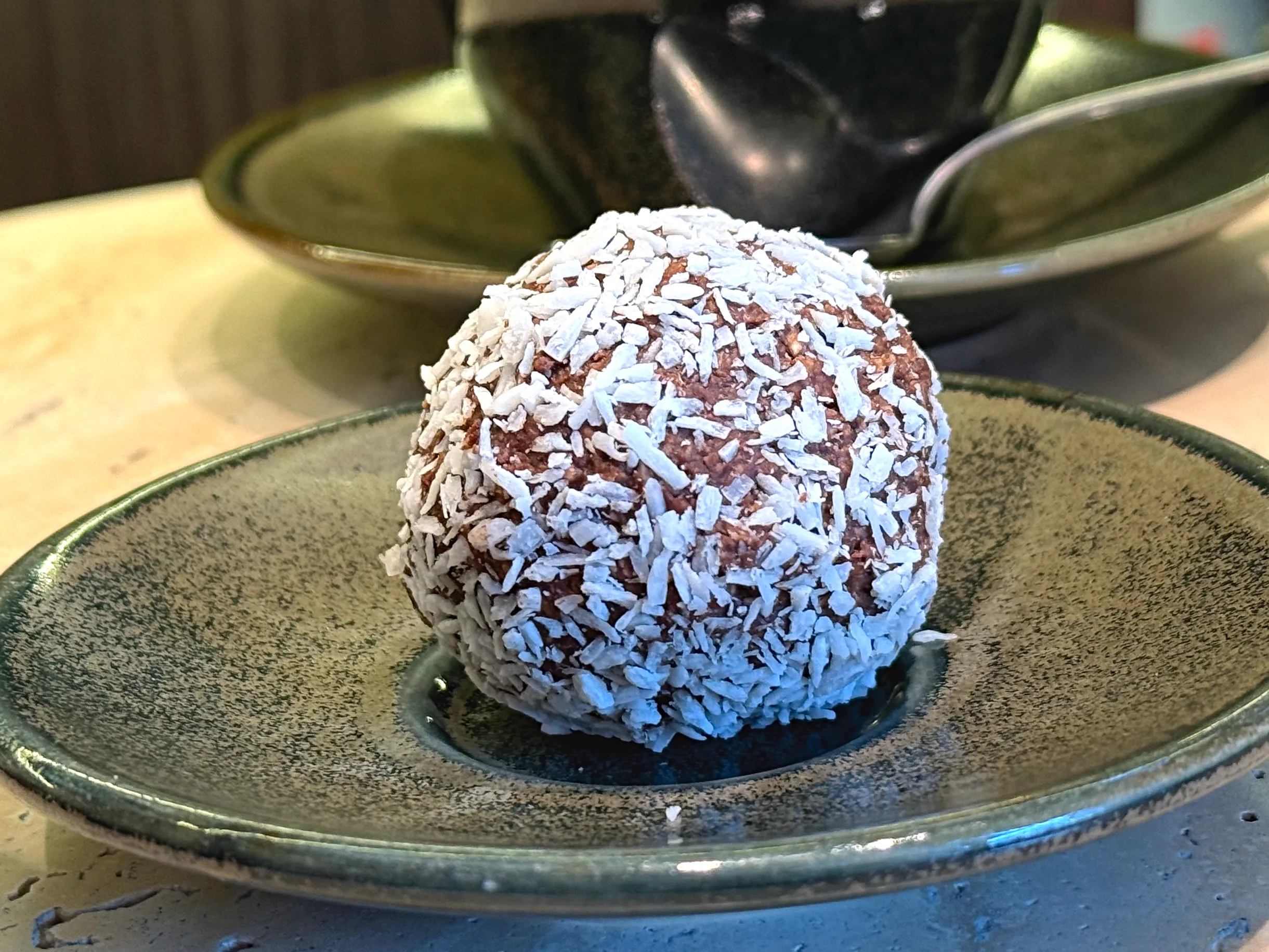 En rund chokladboll helt täckt av kokosflingor på ett litet grönt stengodsfat, med kaffekopp och skålar i bakgrunden.