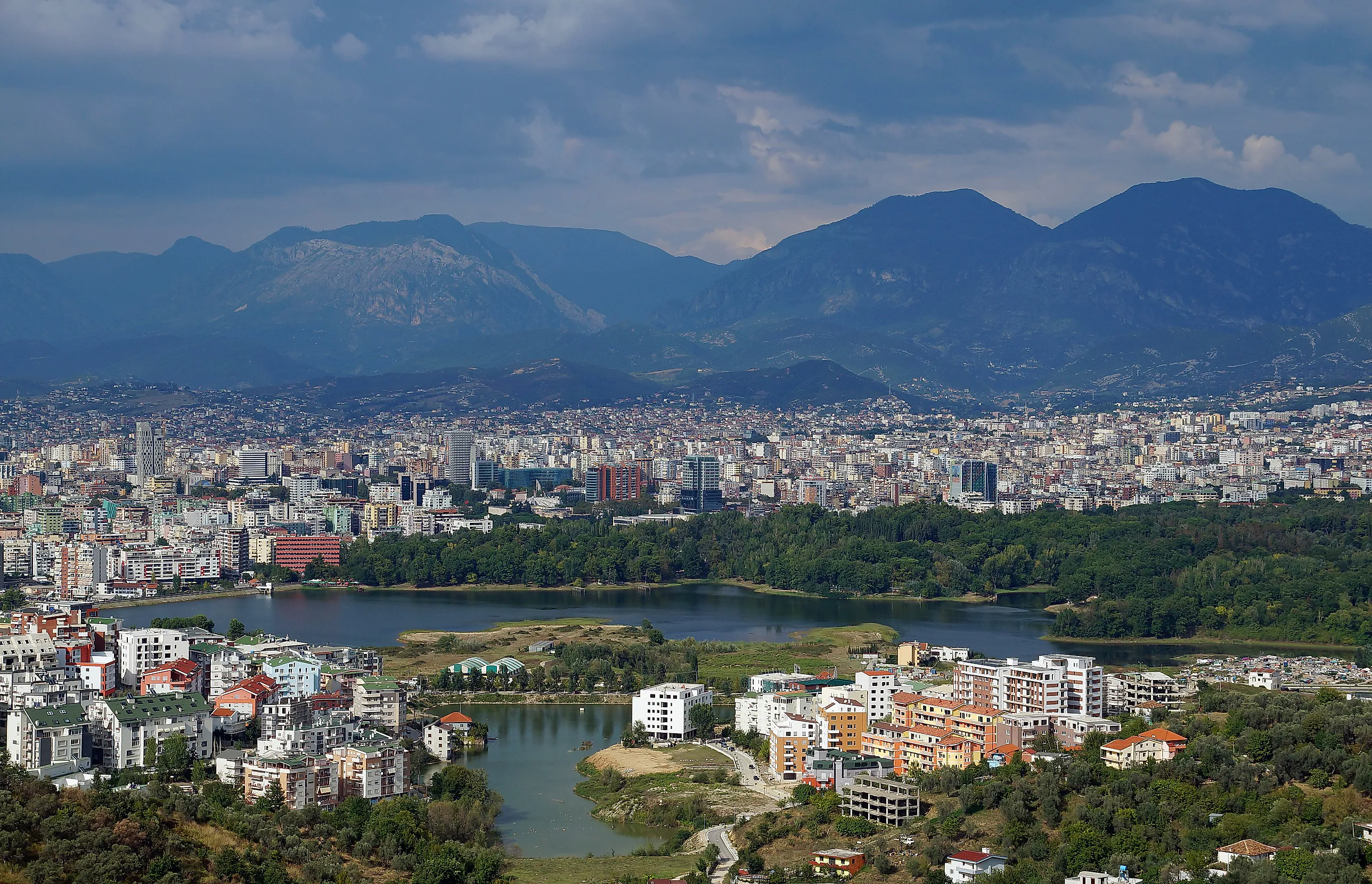 Vidsträckt utsikt över Tirana med tät bebyggelse, en park och en långsmal sjö i förgrunden och blådisiga bergskedjor under molnig himmel i bakgrunden.