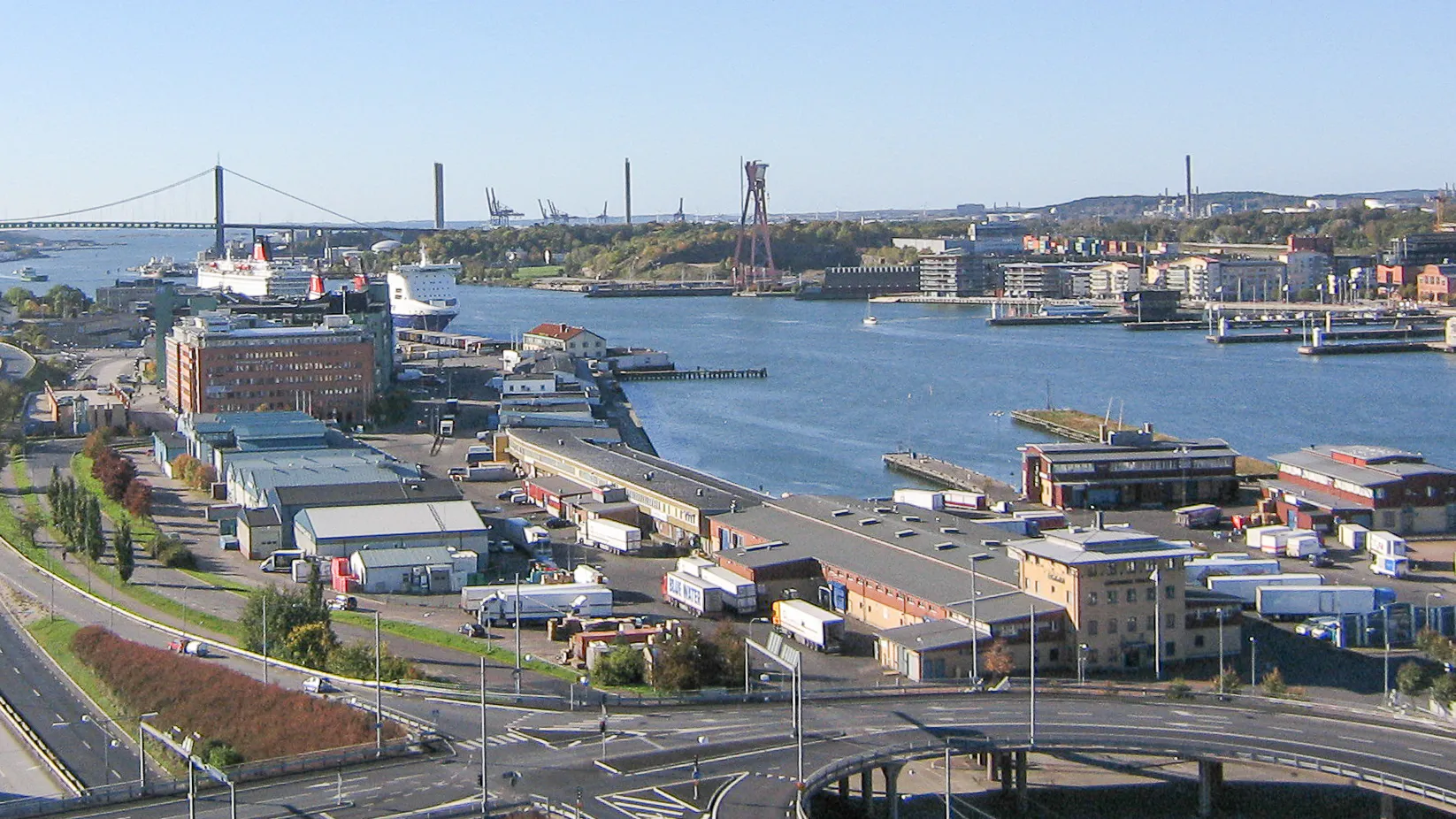 Utsikt över Göta älv i Göteborg med hamnbyggnader, lastbilar och kajer i förgrunden, Älvsborgsbron och en färja i bakgrunden under klarblå himmel.