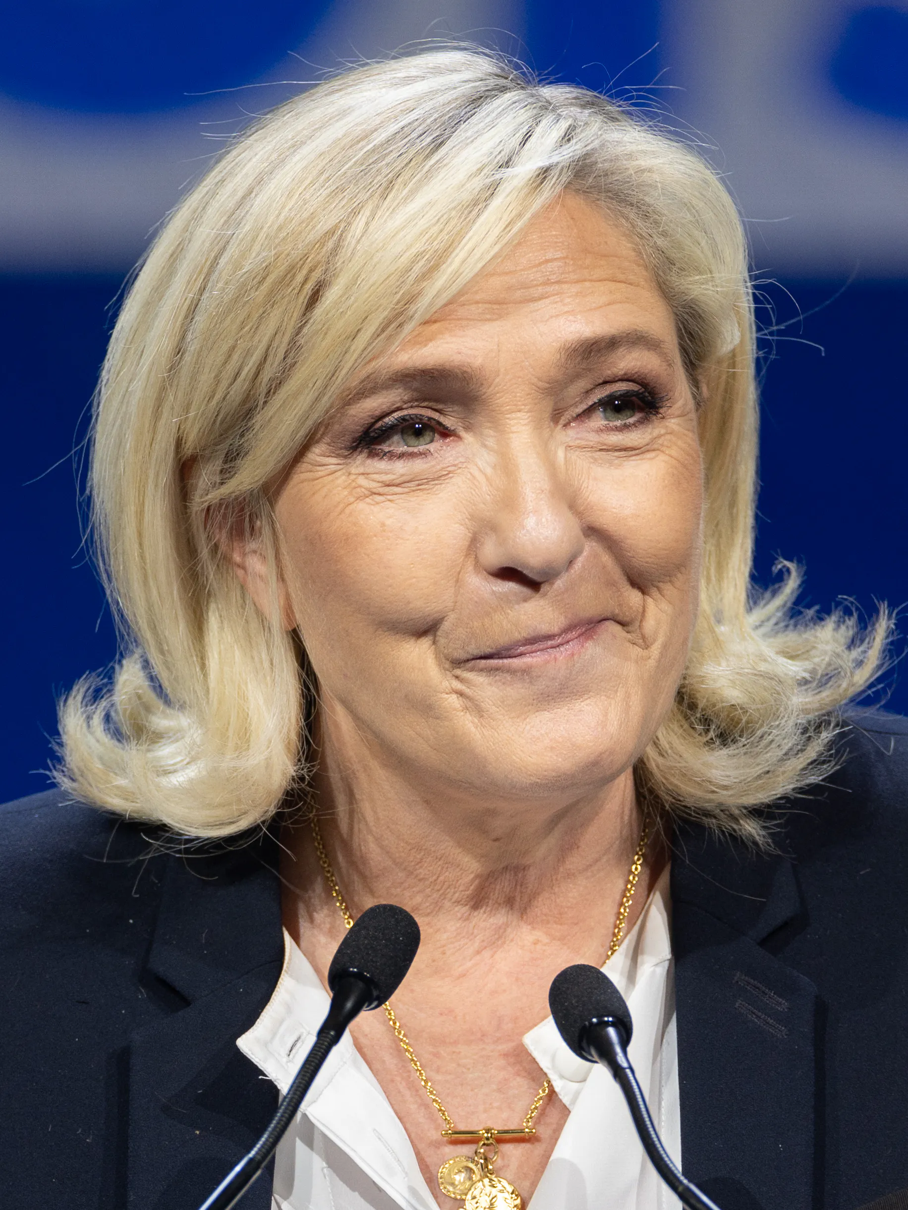 Marine Le Pen i mörk kavaj och vit blus bakom två mikrofoner, med ljust axellångt hår och ett svagt leende.