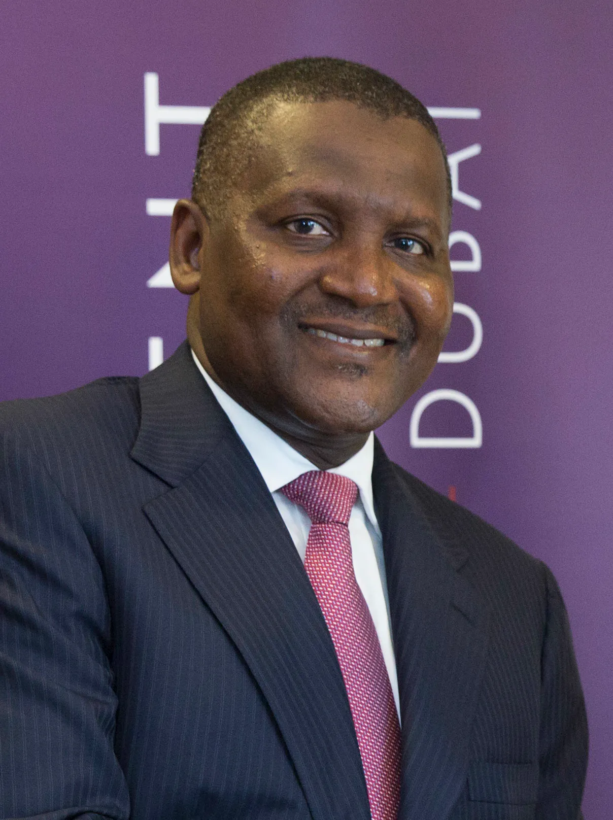 Aliko Dangote i mörk kritstrecksrandig kostym och röd slips, leende framför en lila vägg.