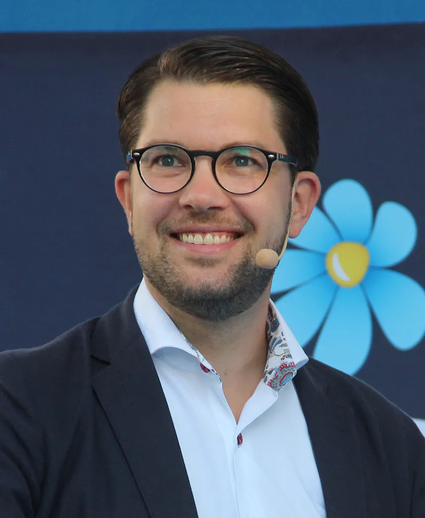 Jimmie Åkesson i mörk kavaj och vit skjorta med myggmikrofon, framför en blå skynke med partiblomman.