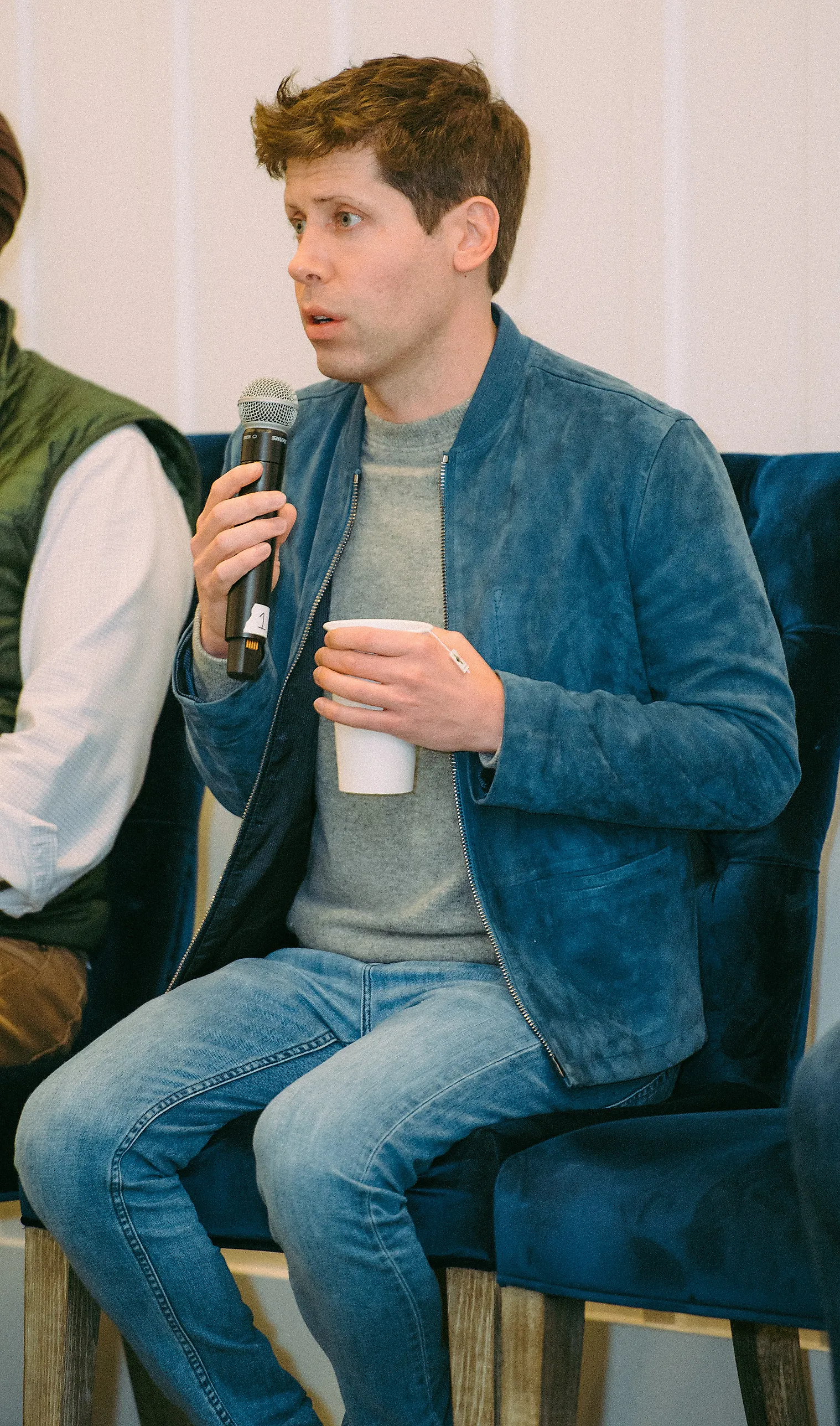 Sam Altman sitter i en fåtölj i blå mockajacka och talar i en handmikrofon med en pappmugg i andra handen.