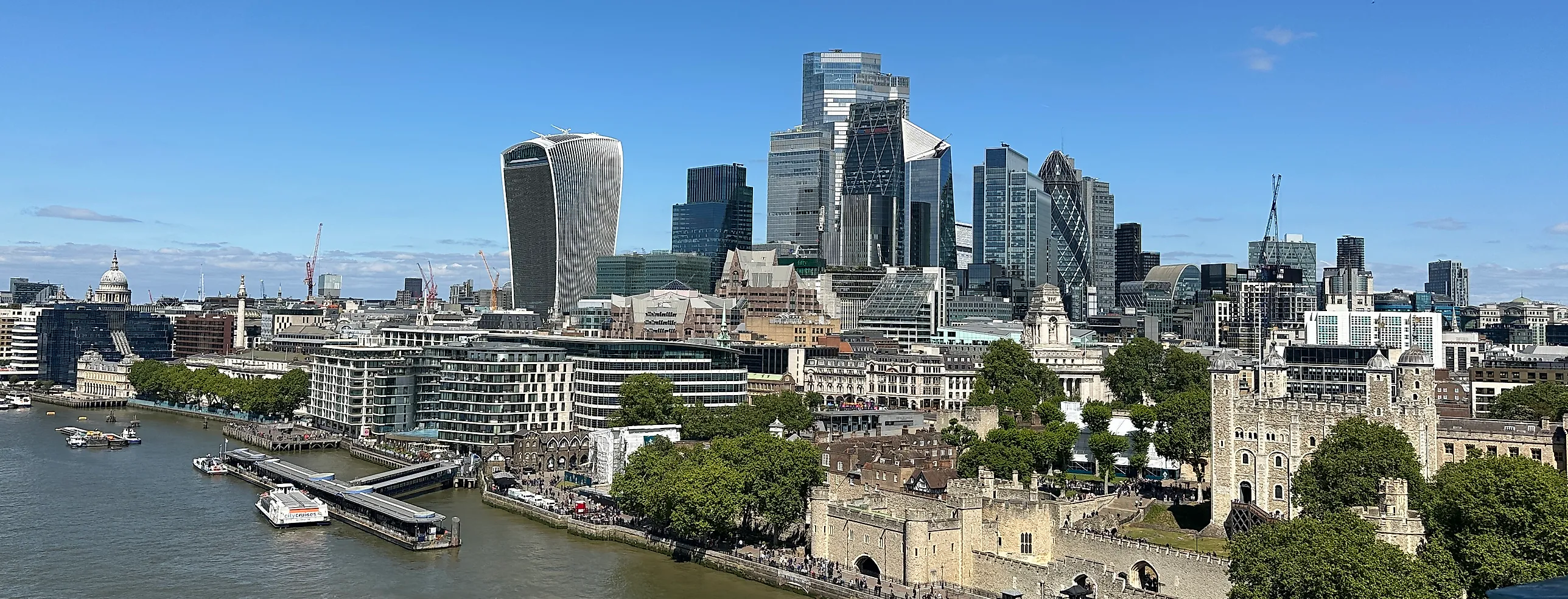 Londons finansdistrikt med glasskyskrapor bakom Themsen och Tower of London i förgrunden, under klarblå himmel.