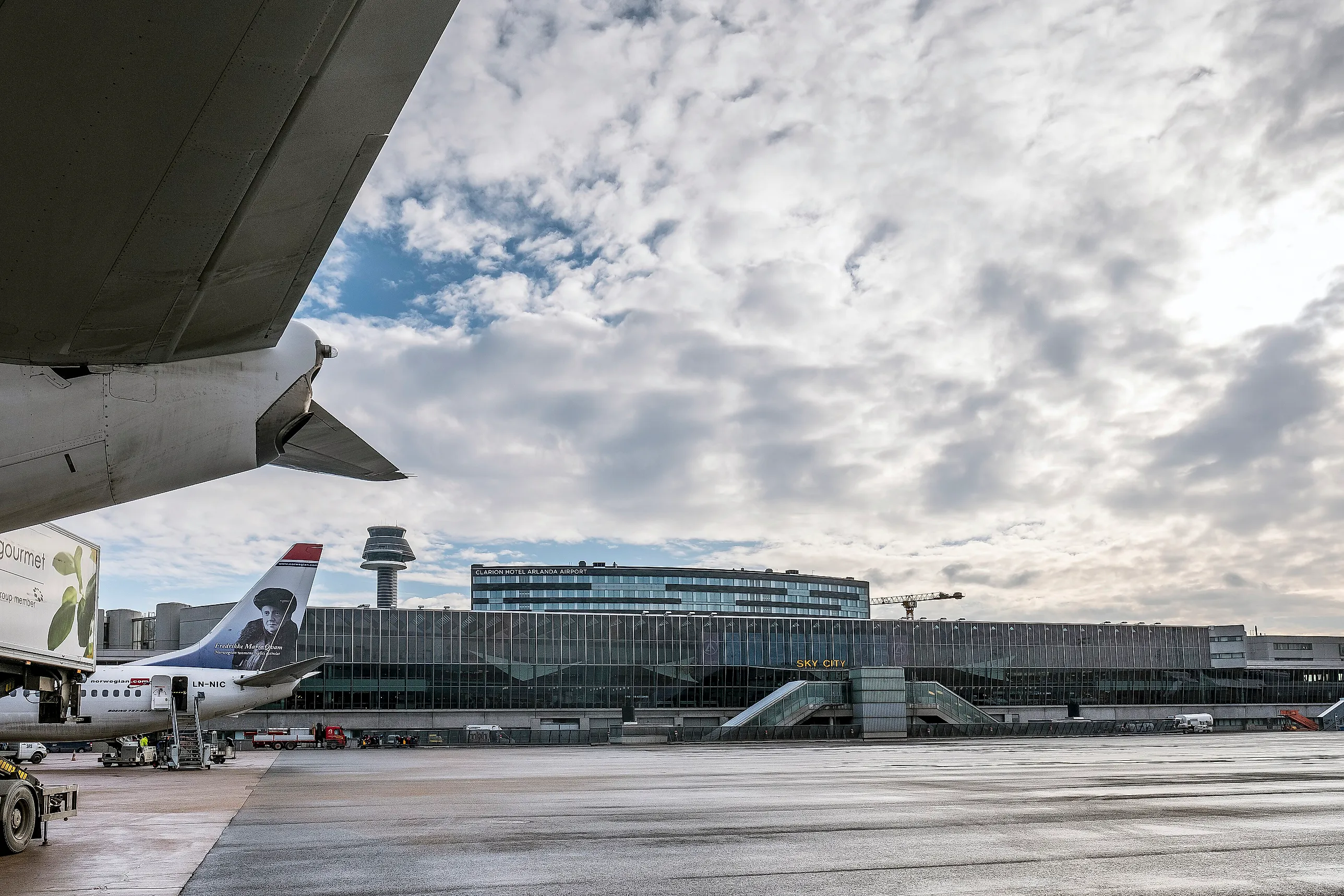 Ett flygplan vid Arlandas platta framför terminalbyggnaden med flygledartornet och hotellet Sky City under molnig himmel.
