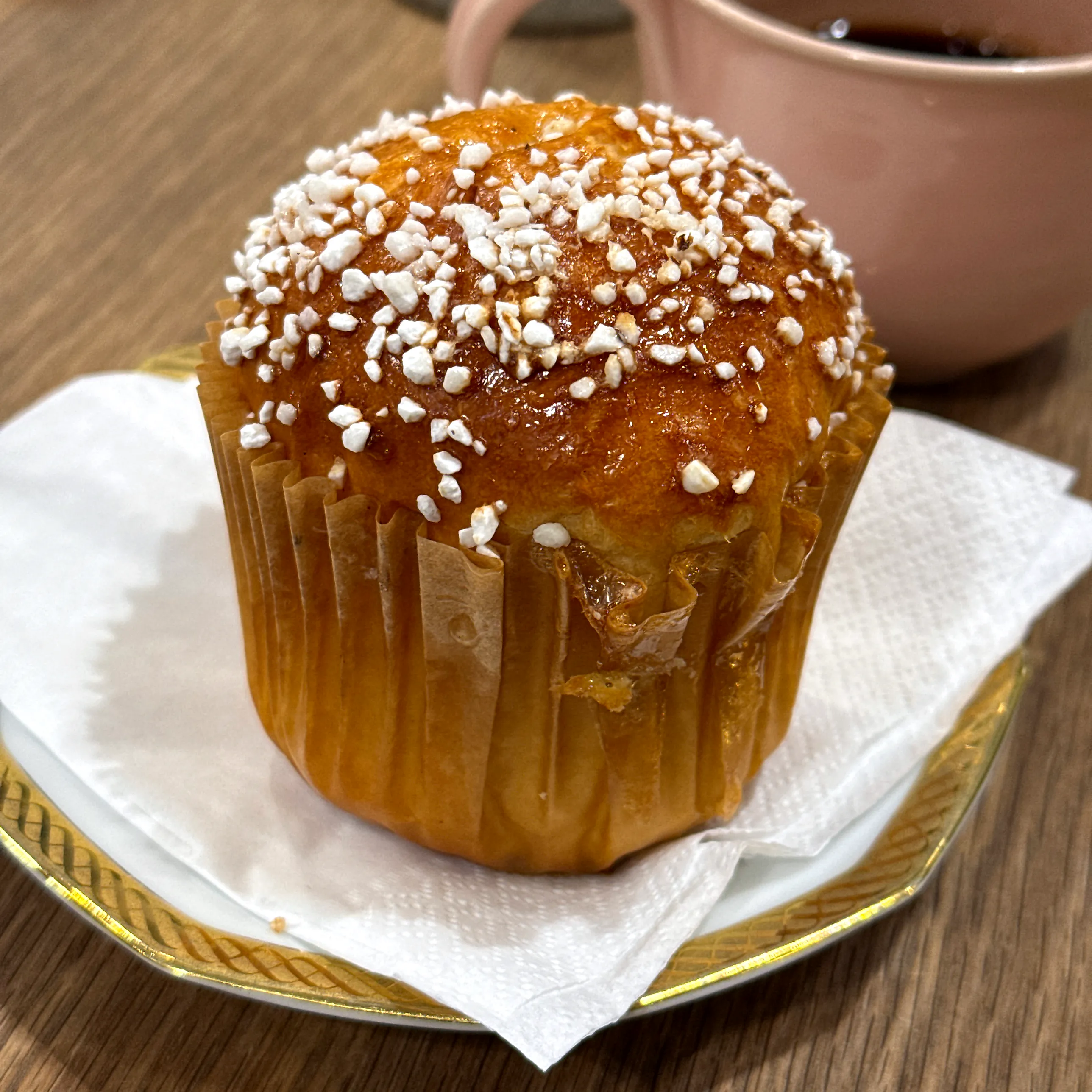 En hög, gyllene vetebulle i brun papperform med pärlsocker på toppen, på en servett och guldkantat fat vid en kaffekopp.