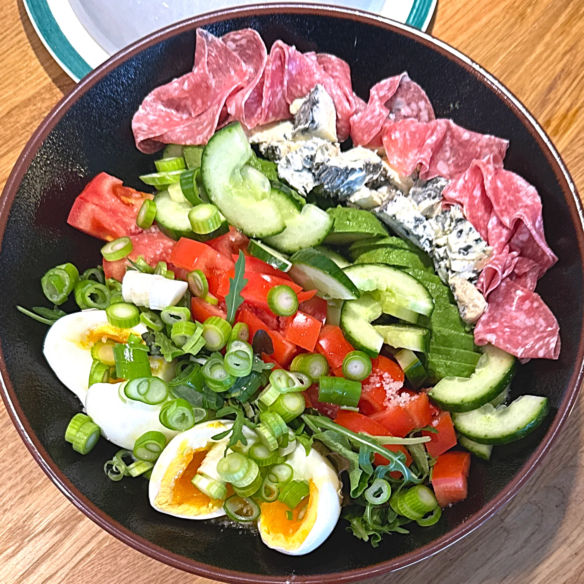 Djup mörk skål med salladsmix toppad i sektioner av tunt skivad salami, smulad ädelost, avokadoskivor, gurka, tomatbitar och halverade ägg med krämig gula, allt överstrött med skivad salladslök.