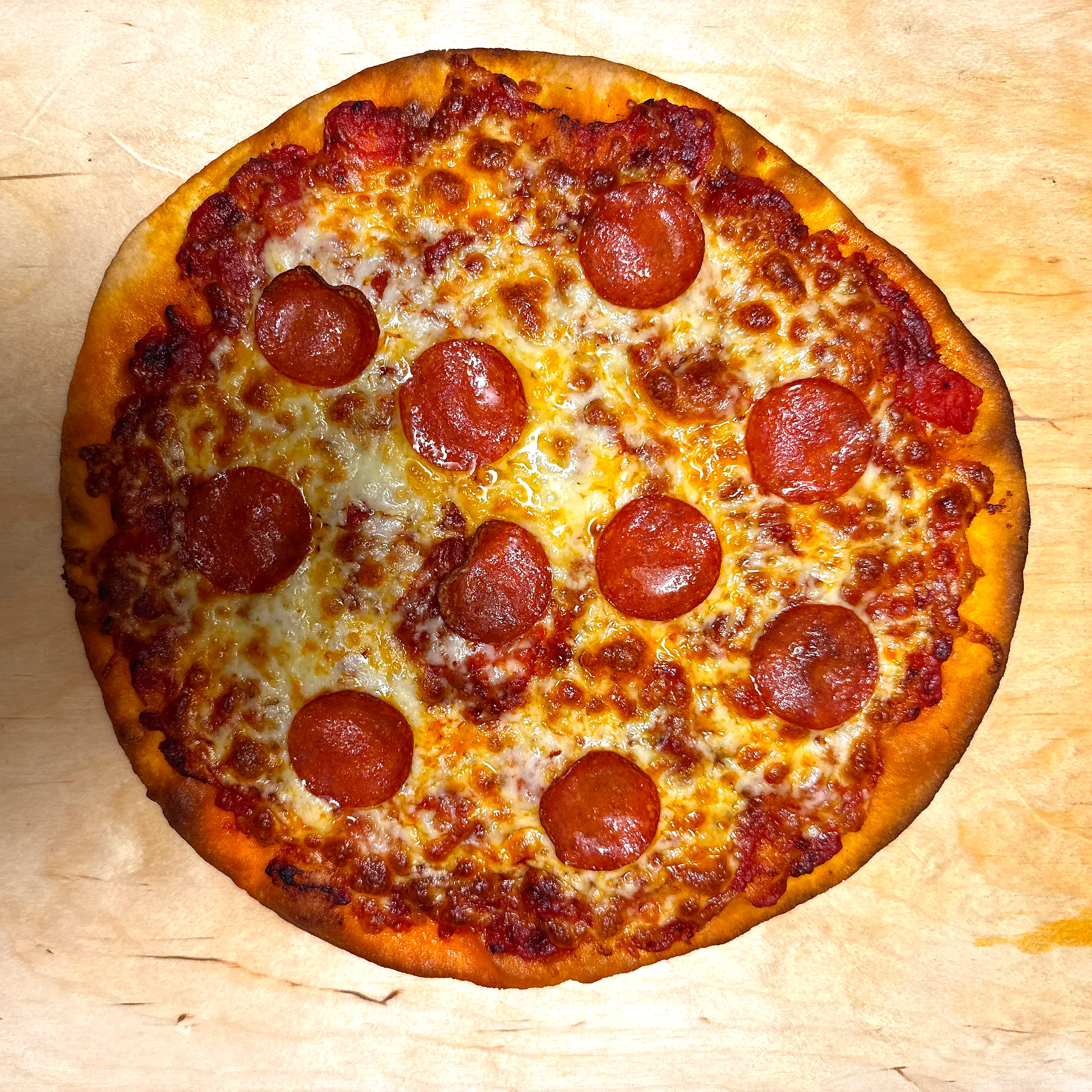 Rund pizza med guldbrun kant, smält ost och tio skivor pepperoni, upplagd på en ljus träskiva.