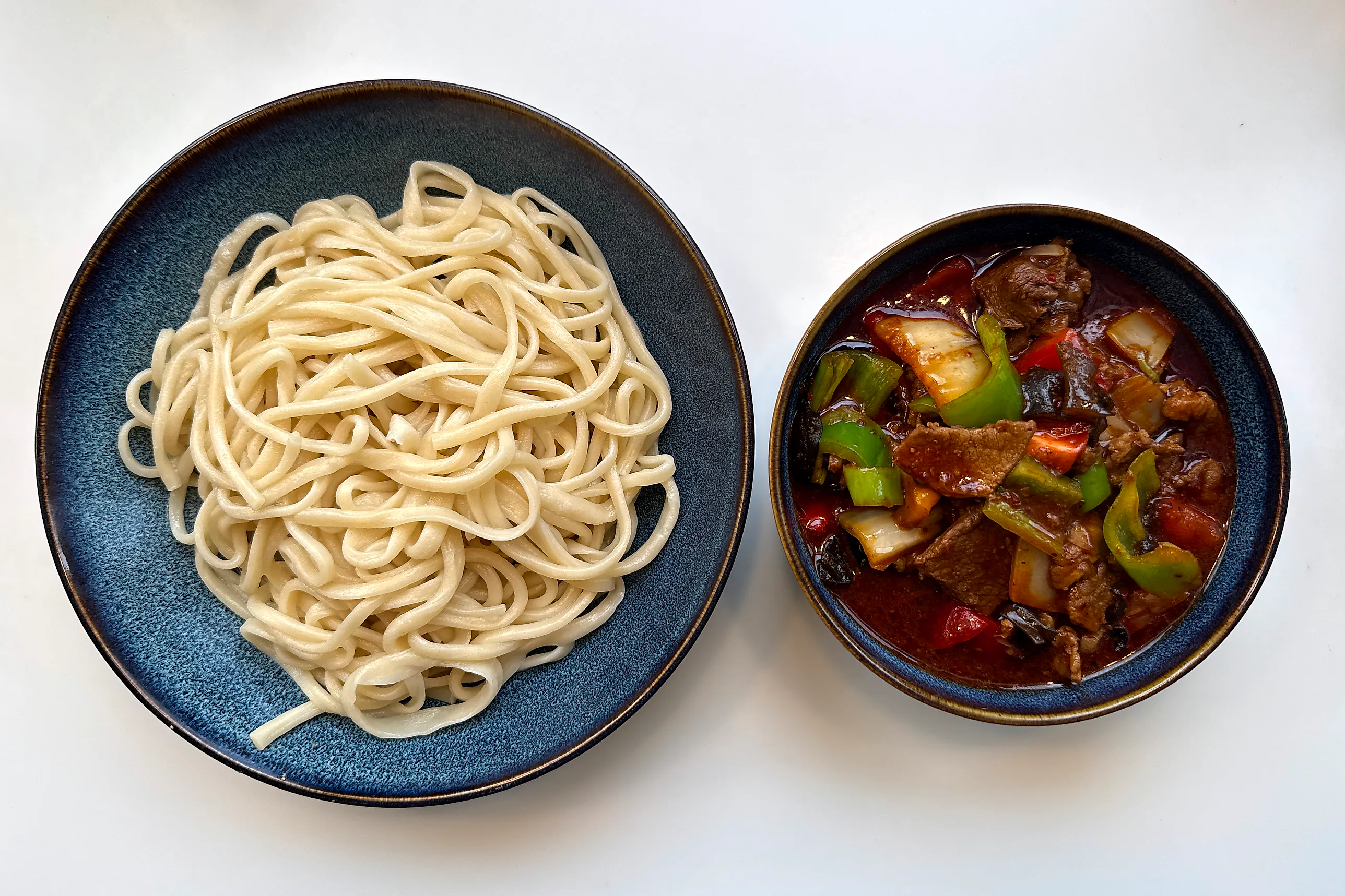 En blå tallrik med en hög ljusa handdragna nudlar och vid sidan en djup skål med mörkröd gryta av lammskivor, grön och röd paprika, lök och svart träsvamp.