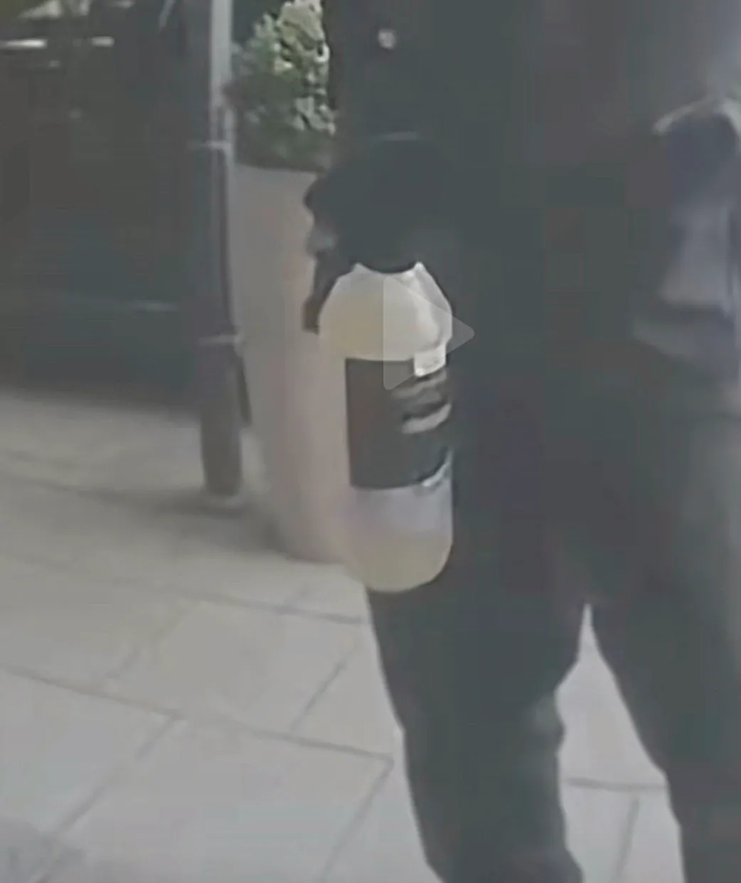 Suddig övervakningsbild av en mörkt klädd person bakifrån på en trottoar, med play-symbol över bilden.