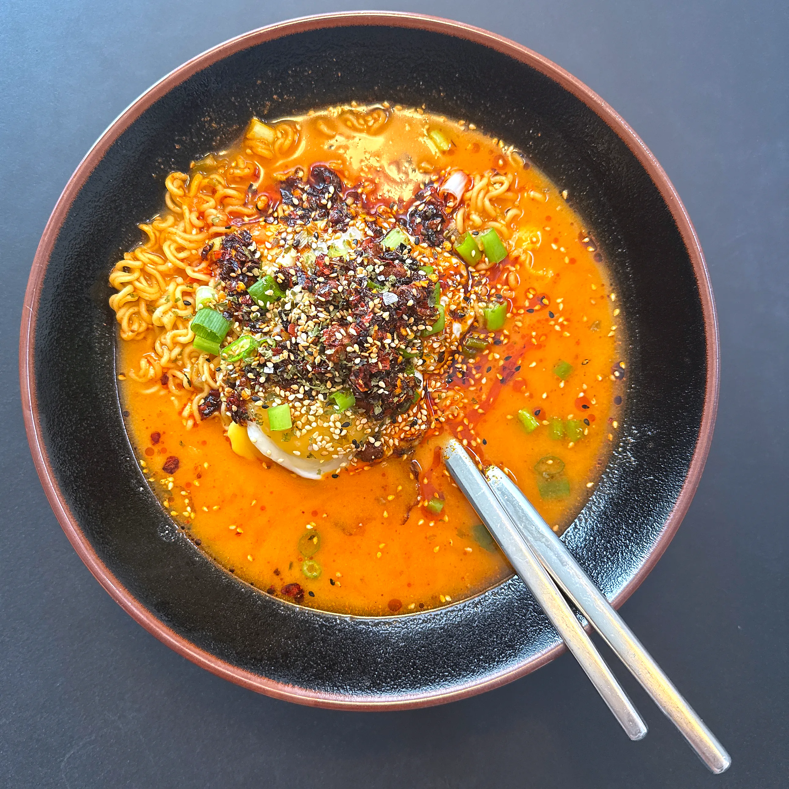 Djup svart skål med snabbnudlar i orangeröd, krämig soppa, ett stekt ägg i mitten under en hög klick crispy chili oil, sesamfrön och strimlad salladslök, med metallpinnar vilande i skålen.