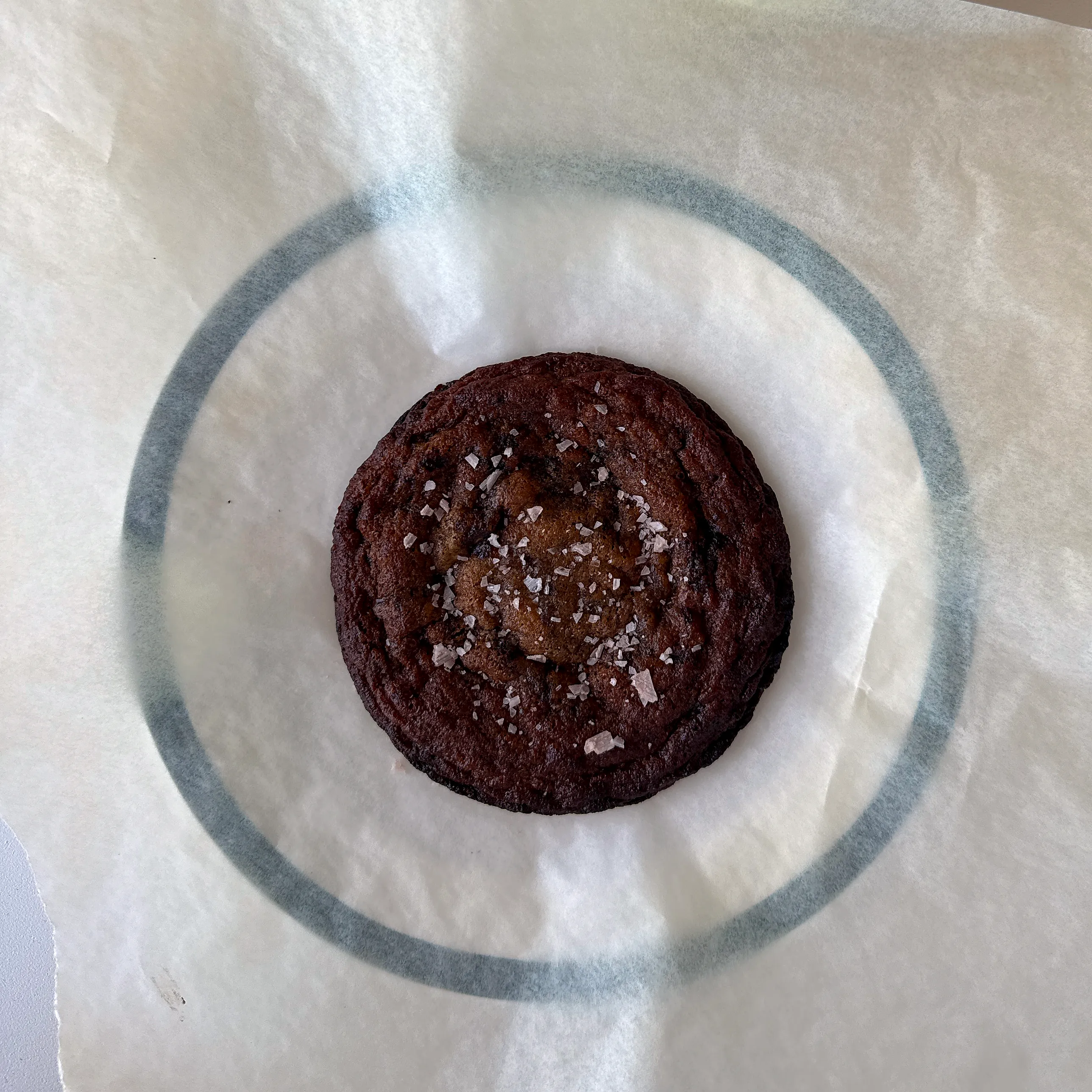 En tjock, mörkt gyllenbrun cookie med skrovlig yta och flingsalt på toppen, liggande på bakplåtspapper.