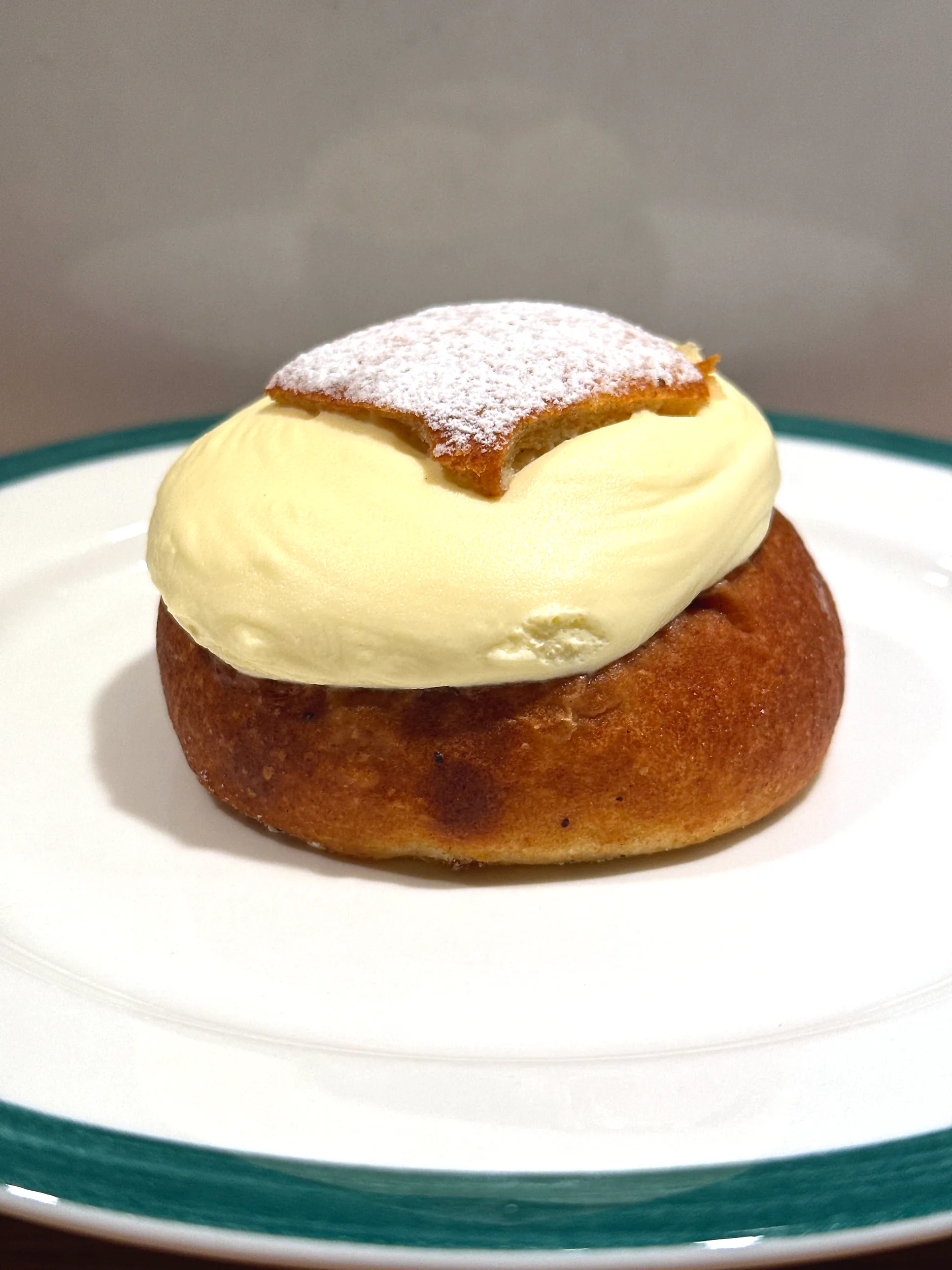 En semla på vit tallrik med grön kant, med hög vispad grädde mellan bullen och det florsockerpudrade locket.