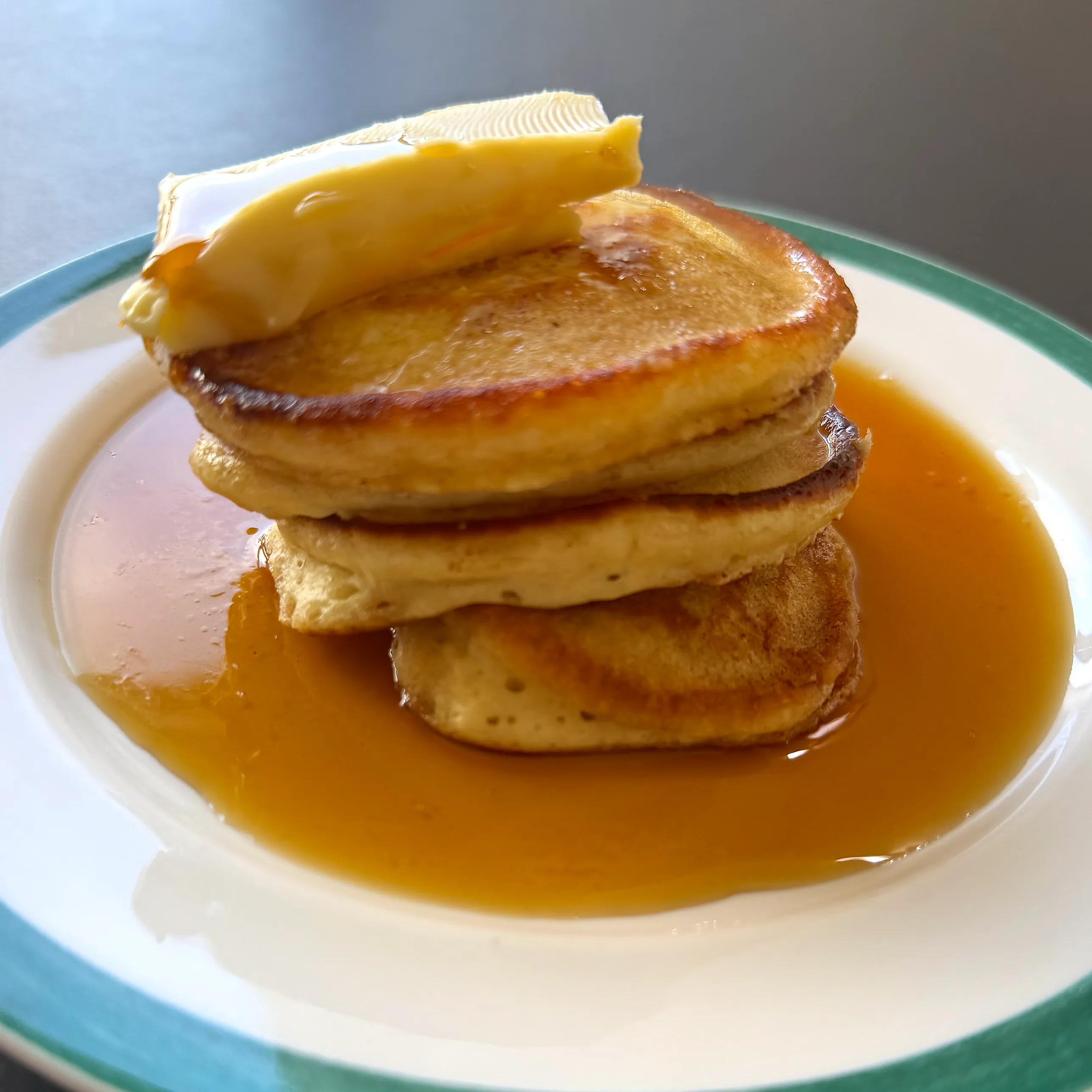 Tre tjocka gyllenbruna pannkakor staplade på en vit tallrik med turkos kant, med en skiva smör på toppen och lönnsirap som runnit ut i en glänsande sjö runt stapeln.