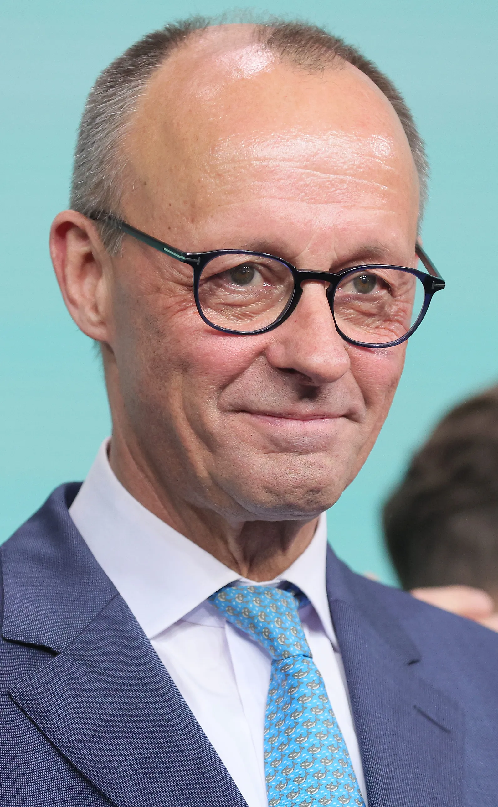 Friedrich Merz i grå kavaj, vit skjorta och ljusblå slips, med mörka glasögon, mot en turkos bakgrund.