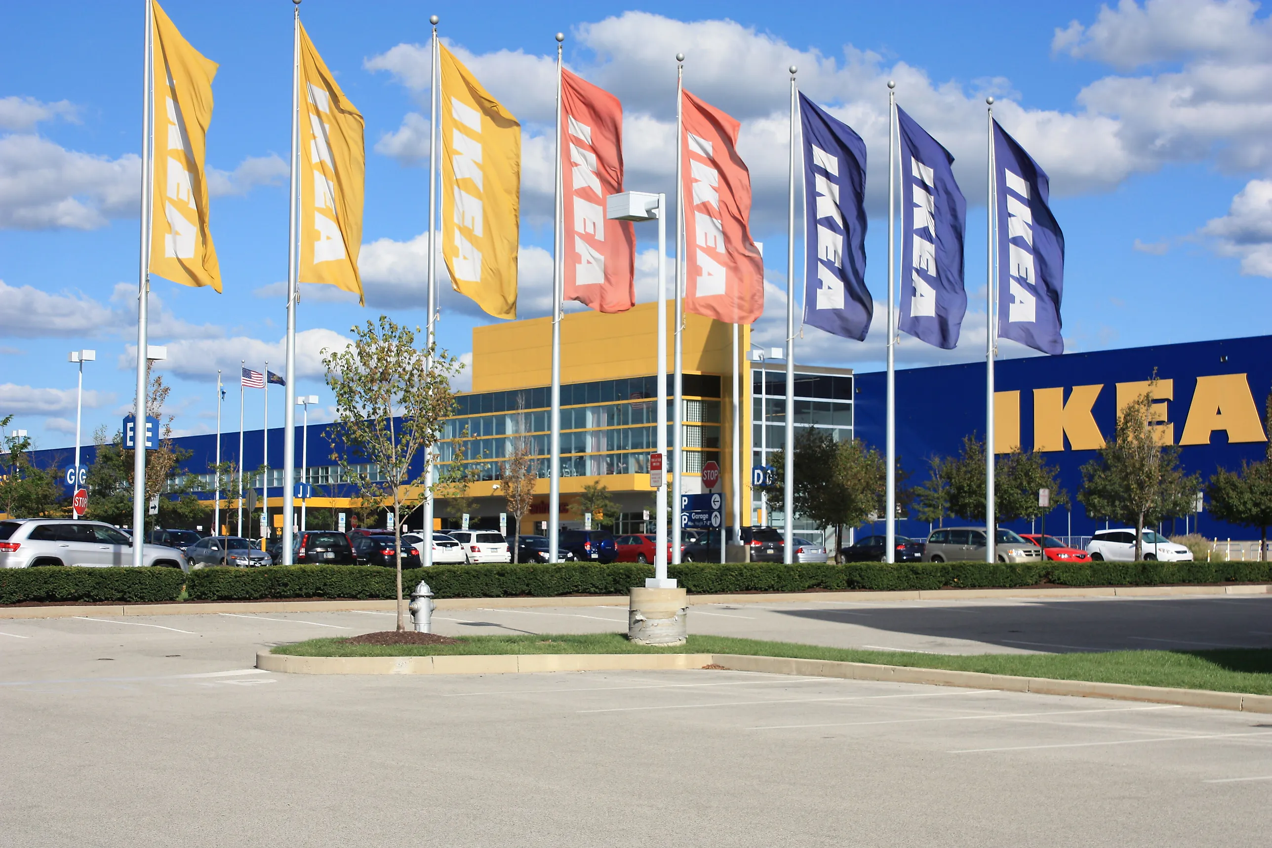 Blågult Ikea-varuhus bakom en rad vajande flaggor med Ikeas namn, med parkeringsplats i förgrunden.