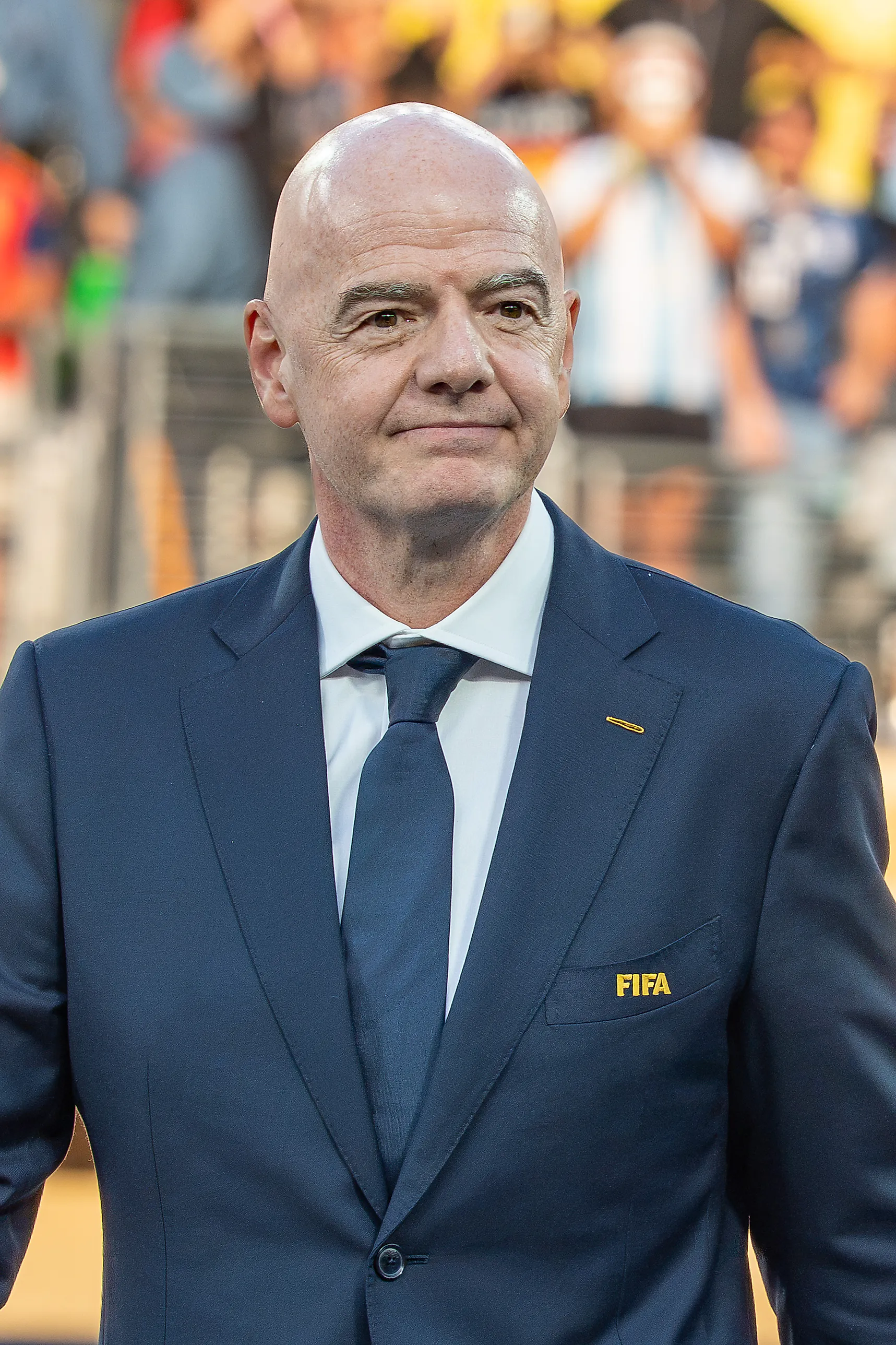 Skallig man i mörkblå kostym med Fifa-märke på bröstfickan står på en fotbollsarena.