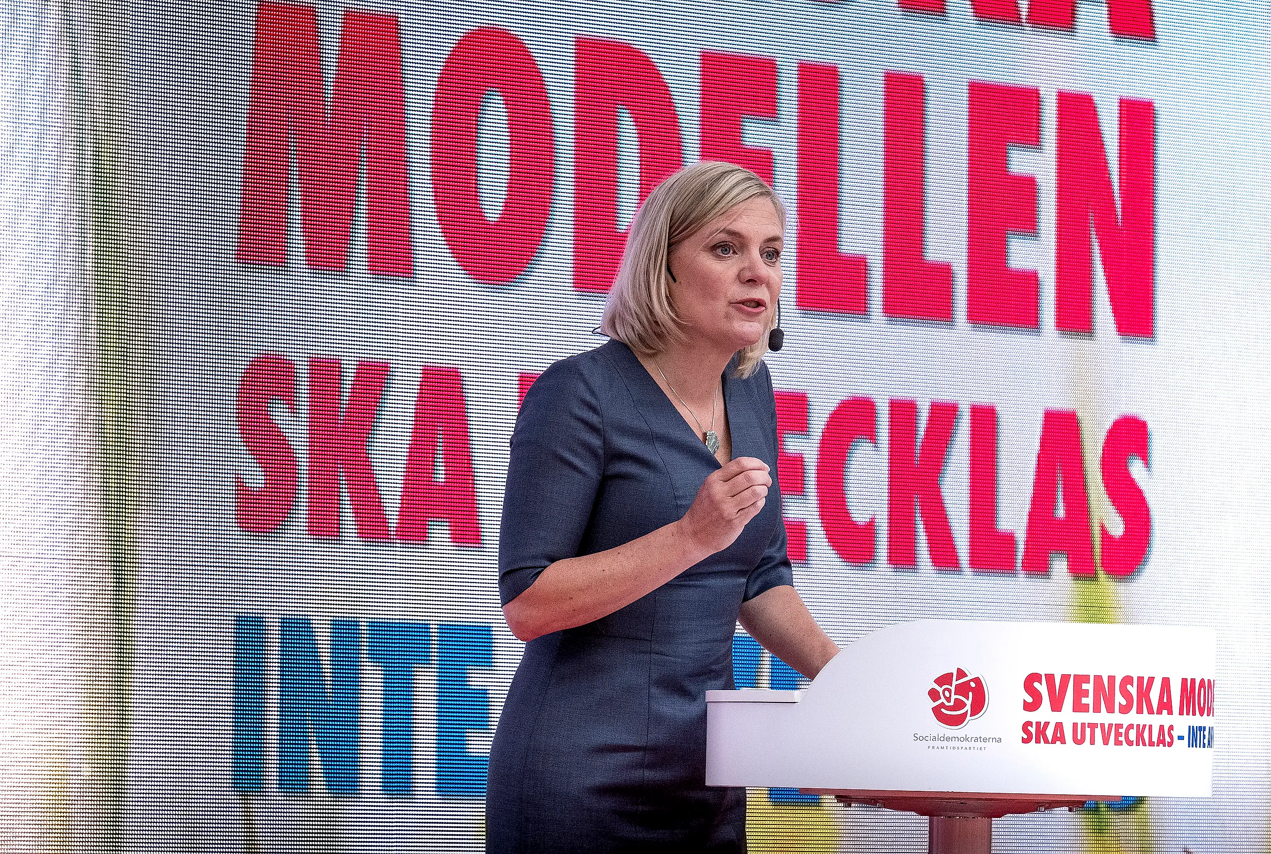 Magdalena Andersson står vid ett talarpodium med mikrofon vid munnen framför en stor skärm med röd text.