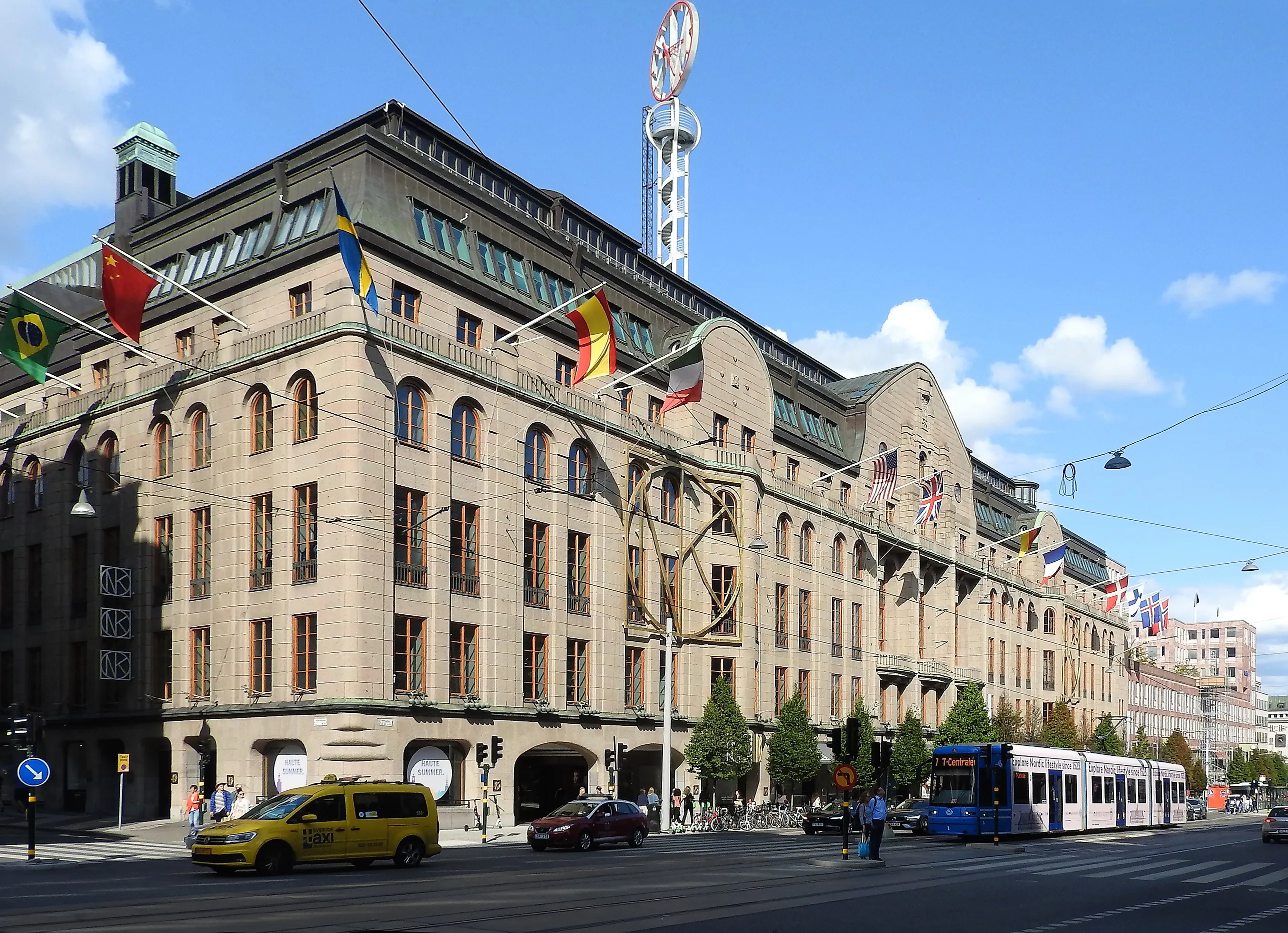 NK:s stora stenvaruhus i hörnet vid Hamngatan i Stockholm med flaggor på fasaden, klocktornet på taket och en spårvagn som passerar.