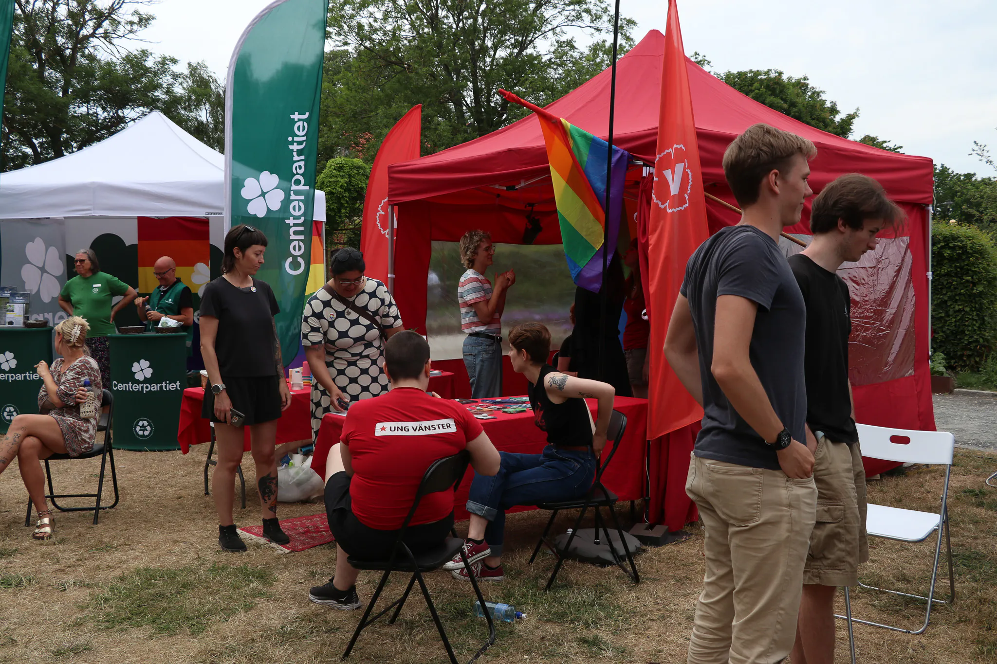 Ett rött tält med Vänsterpartiets V-logotyp och en regnbågsflagga i Almedalen, med besökare framför och Centerpartiets gröna tält intill.