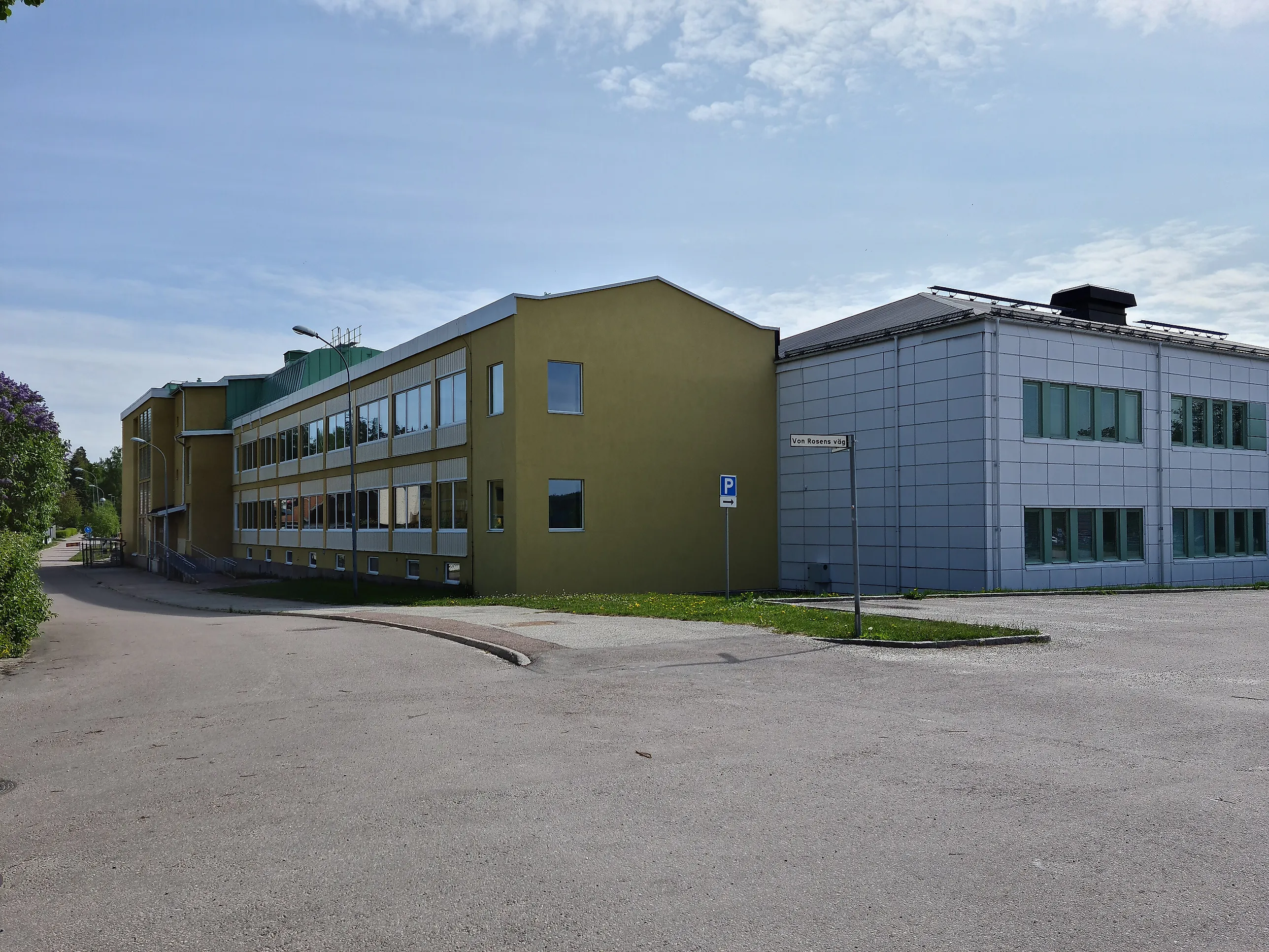Skolbyggnad i två våningar med gulgrön puts intill en ljusgrå byggnadsdel, sedd från en tom asfalterad parkering en solig dag.