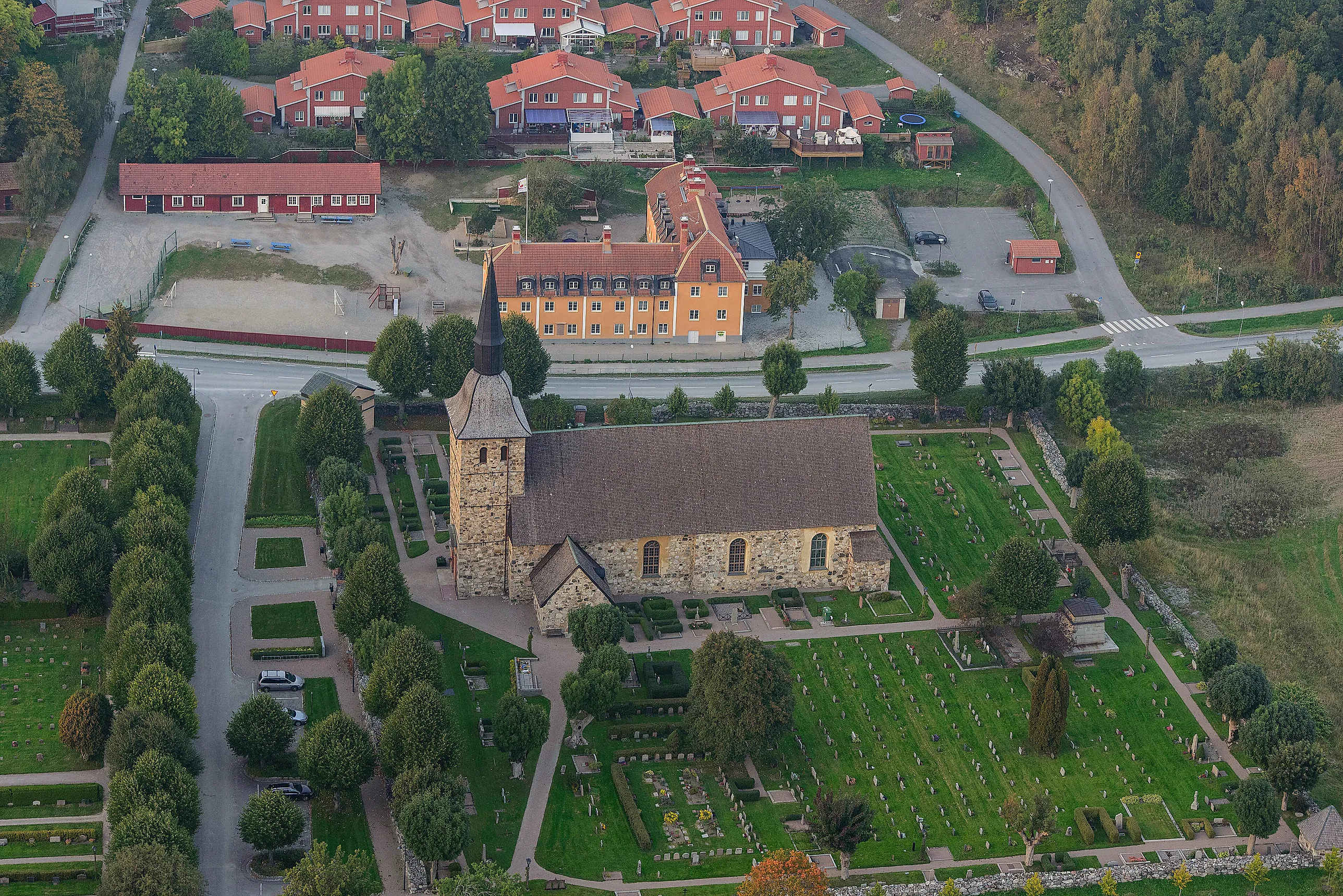 Flygbild över en medeltida stenkyrka med kyrkogård, omgiven av villor och röda hus i Botkyrka.