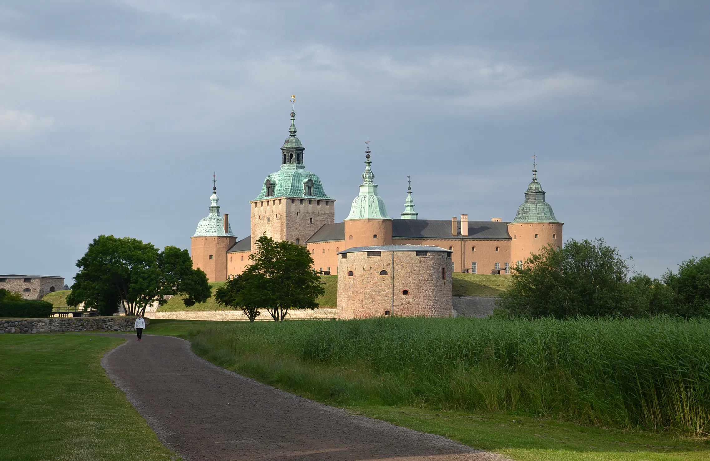Kalmar slott med sina gröna spiror bakom en grusgång och vass en molnig sommardag.