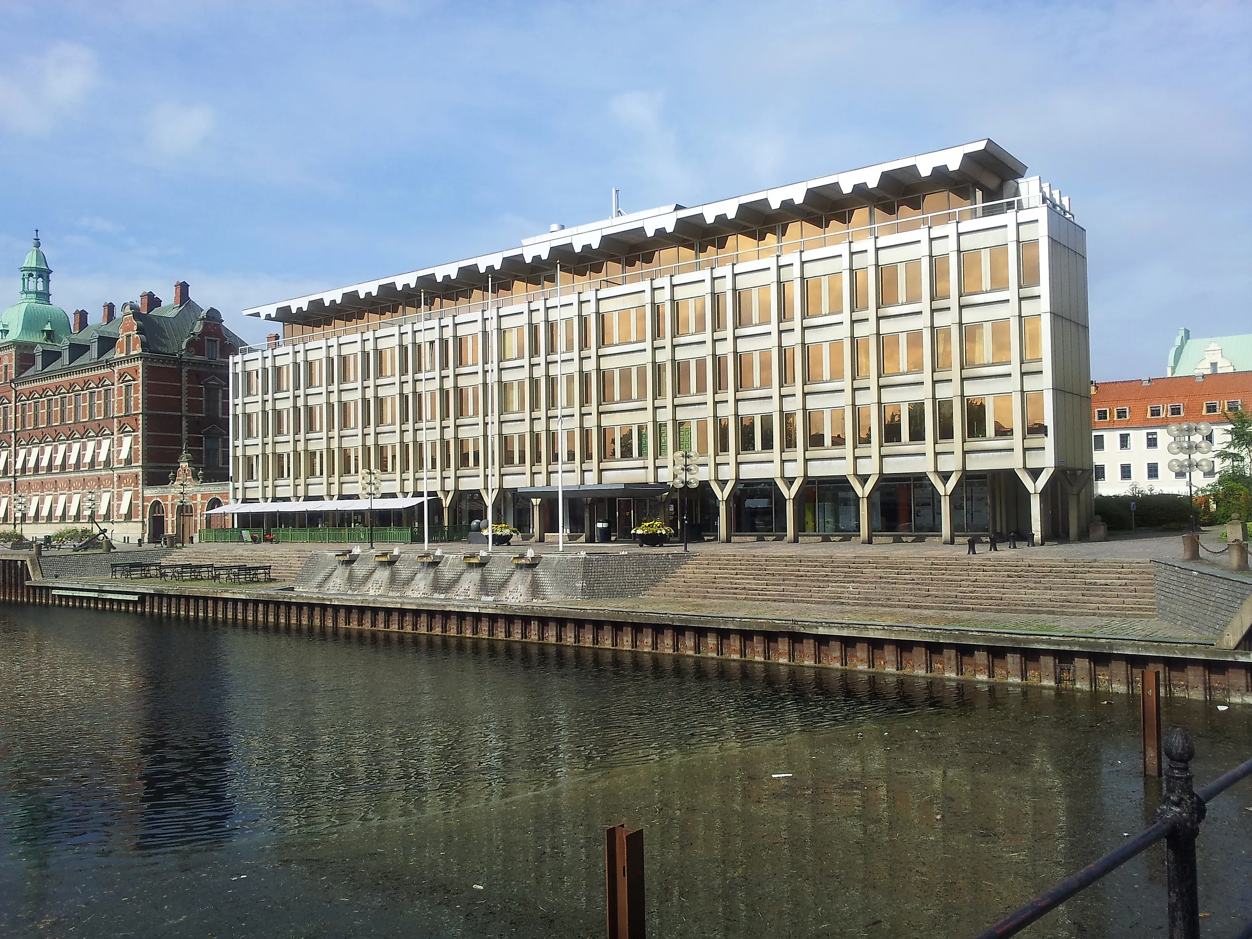 Ett modernt, ljust kontorshus i flera våningar vid en vattenkanal, med trappor ner mot vattnet och äldre tegelbyggnader intill.