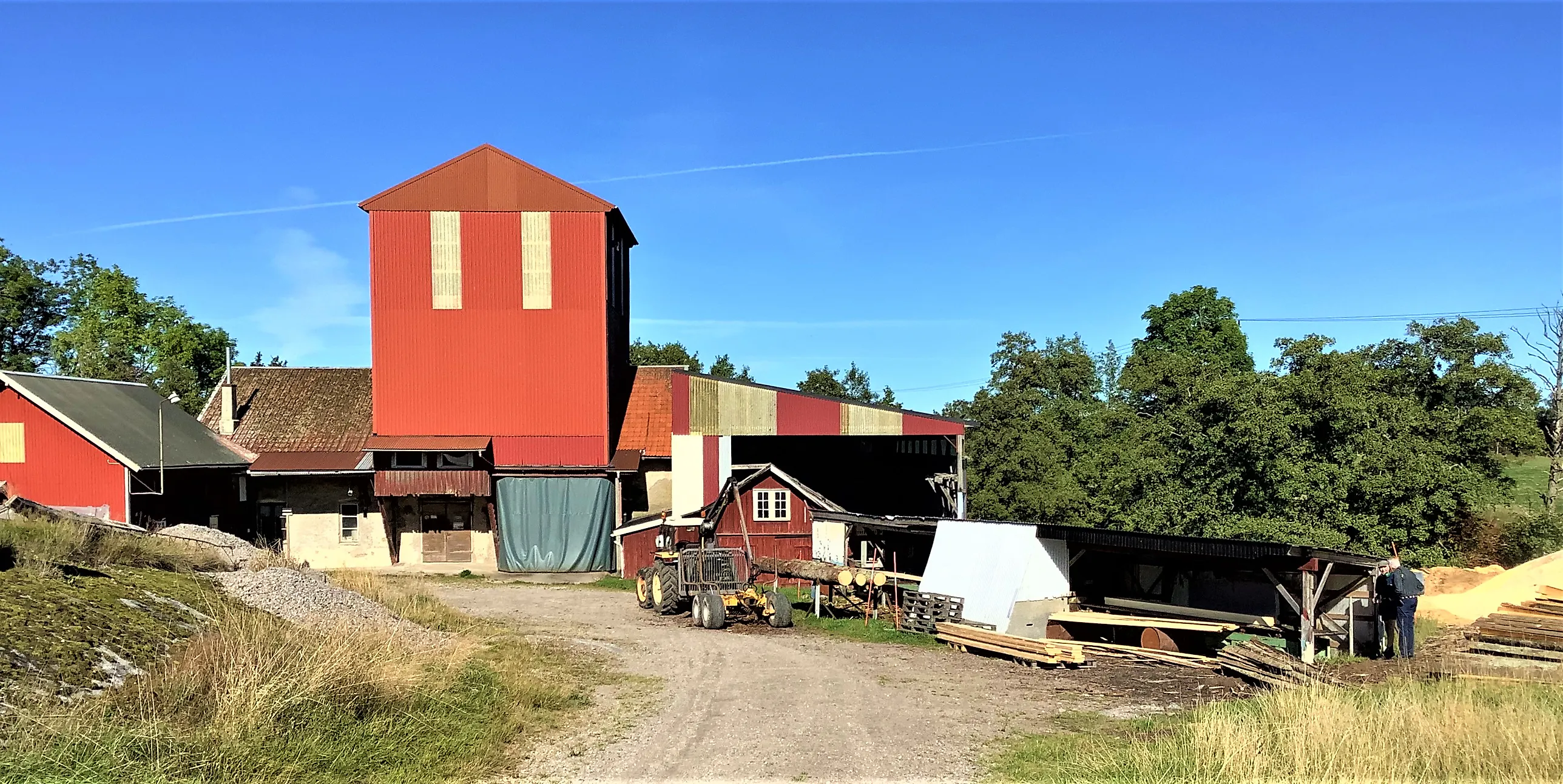 Röd industribyggnad i flera plan med intilliggande sågskjul och grusväg, omgiven av träd under blå himmel.