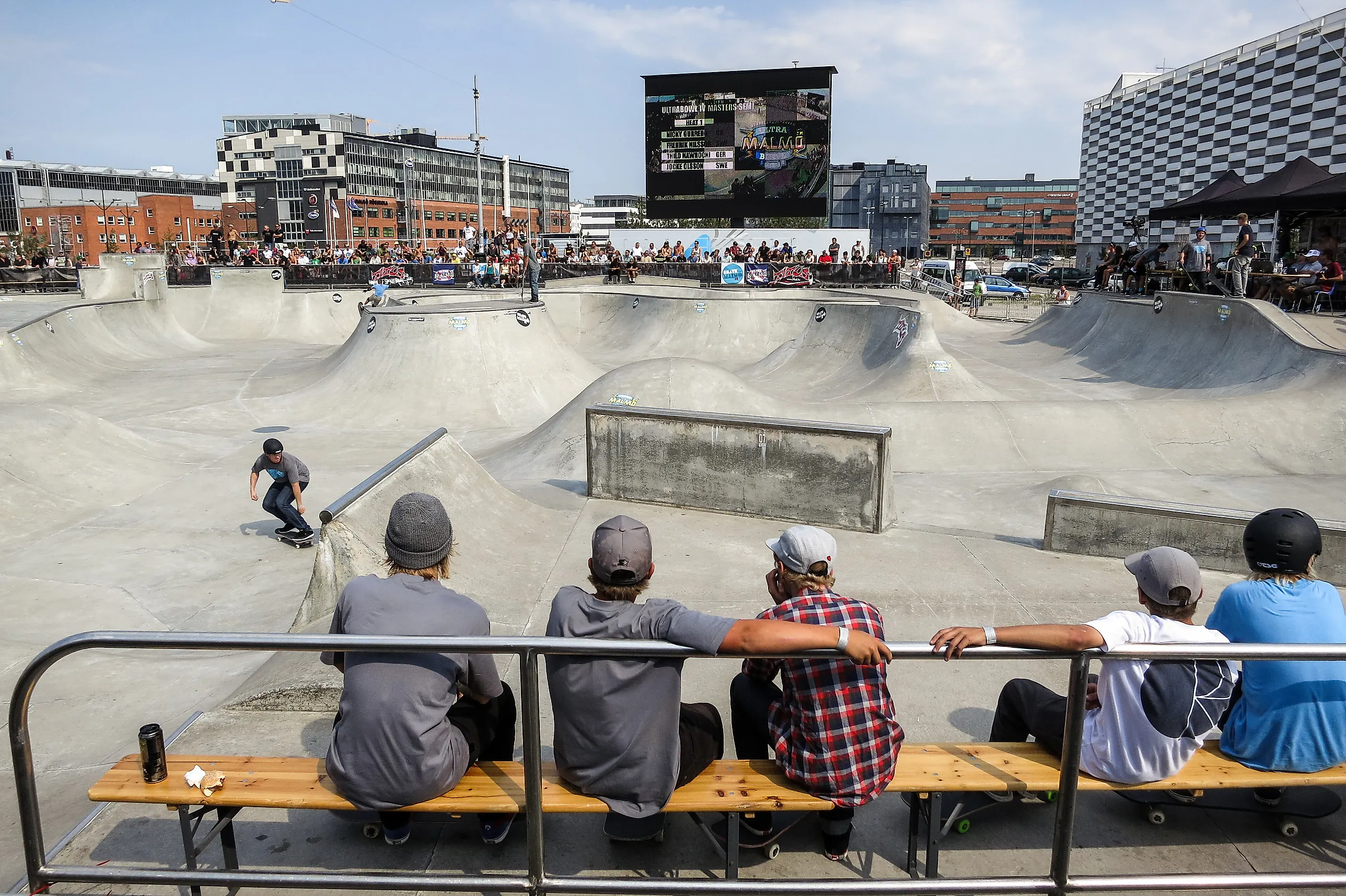 En skejtare åker i en betongbowl i Stapelbäddsparken medan flera personer sitter på en bänk och tittar på, med Västra hamnens byggnader i bakgrunden.
